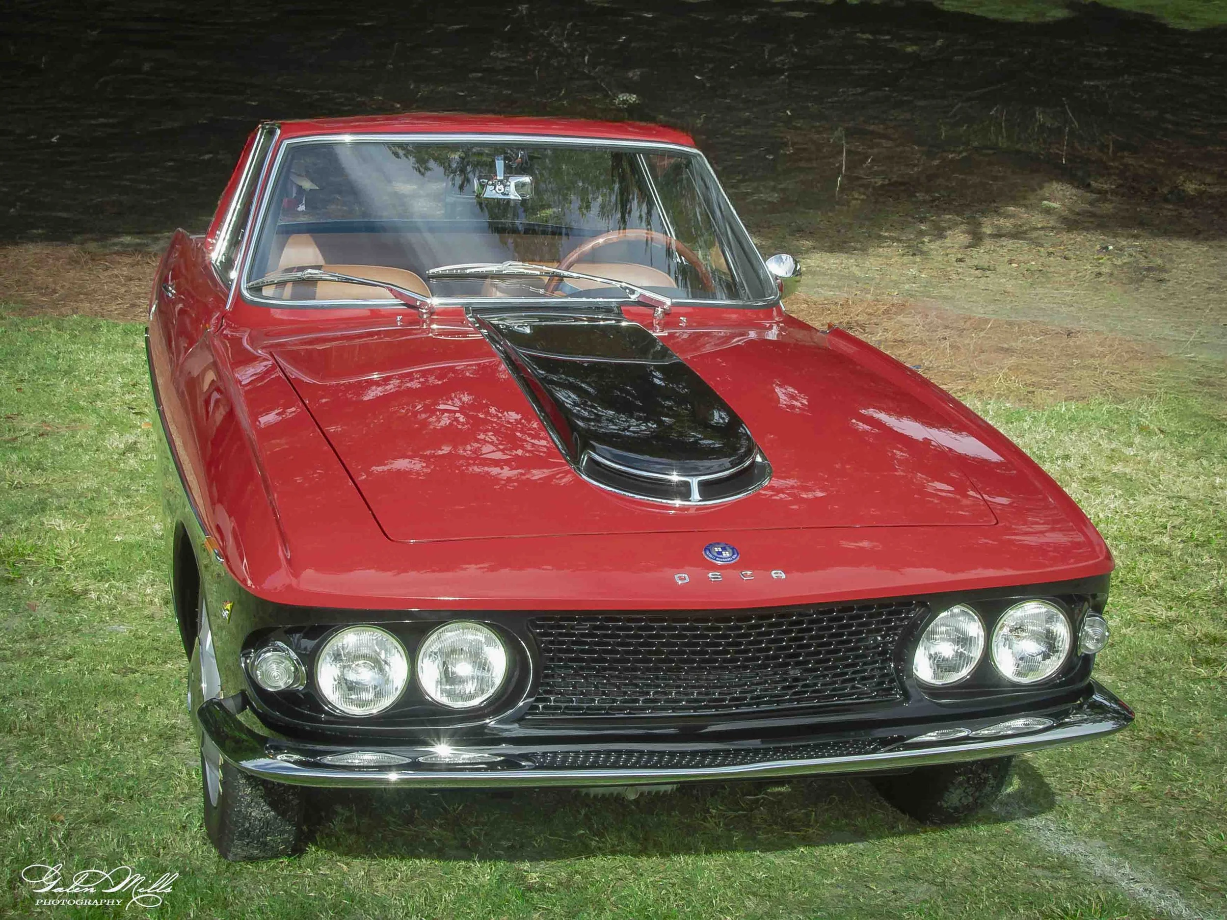 Front view of a classic red sports car with chrome accents and quad headlights, parked on grass with trees in the background.