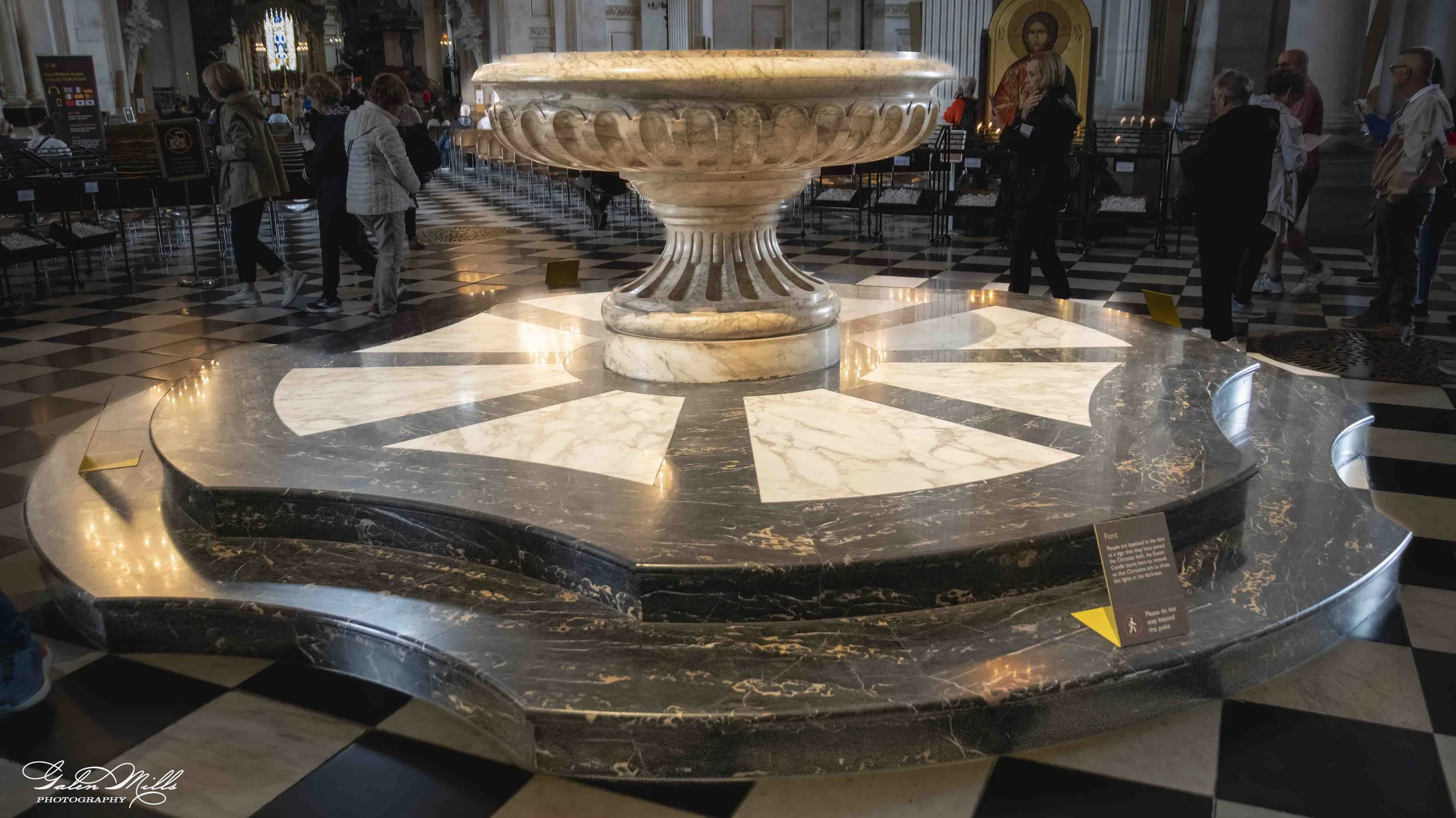 A large ornate marble baptismal font inside a church, surrounded by visitors. The font is elevated on a circular base, with a wide bowl on top, and features carved decorative details. The floor beneath it has a black and white geometric tile pattern.