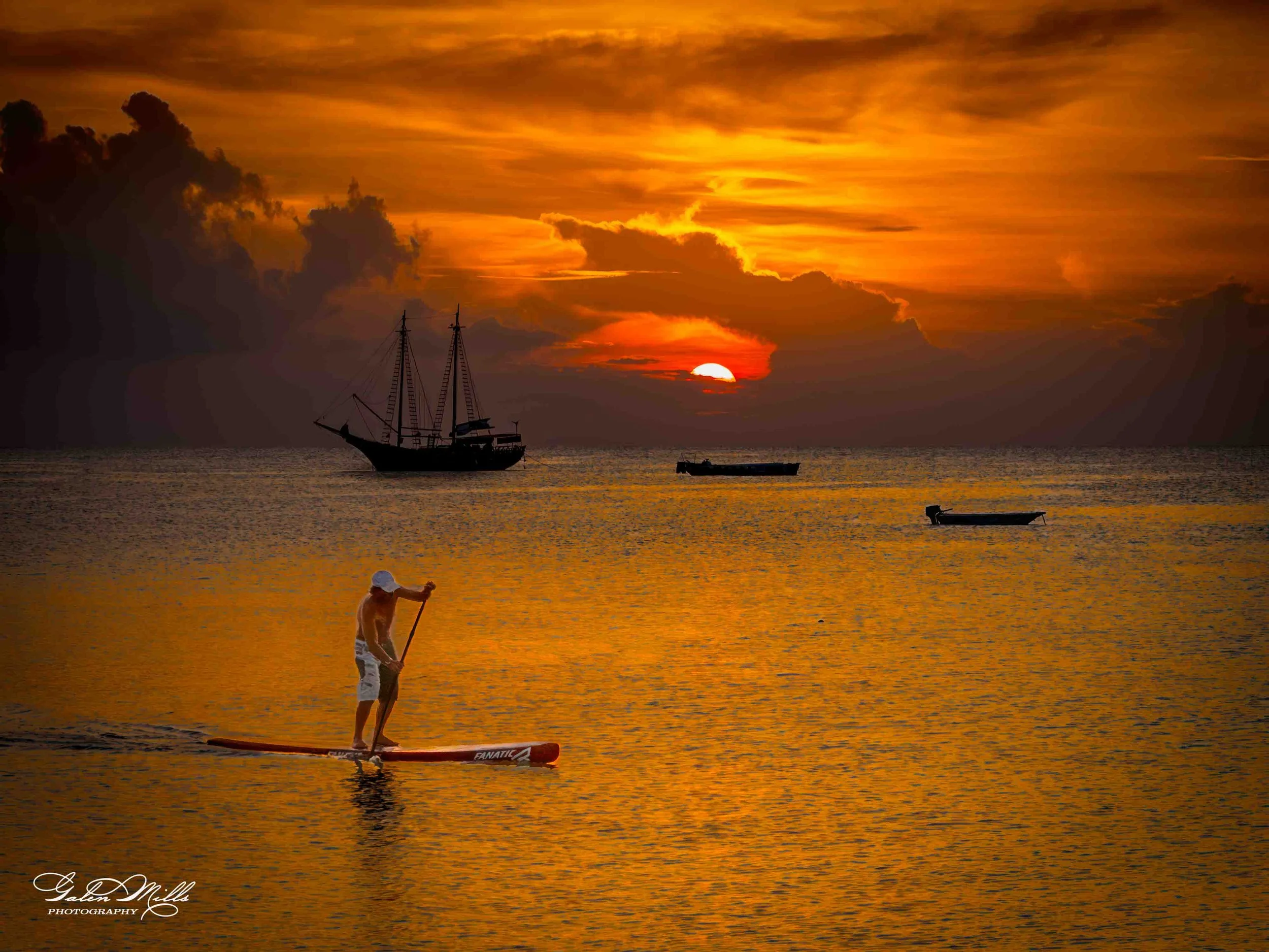 A person paddleboarding on calm water during a sunset, with a large sailing ship and smaller boats in the background.