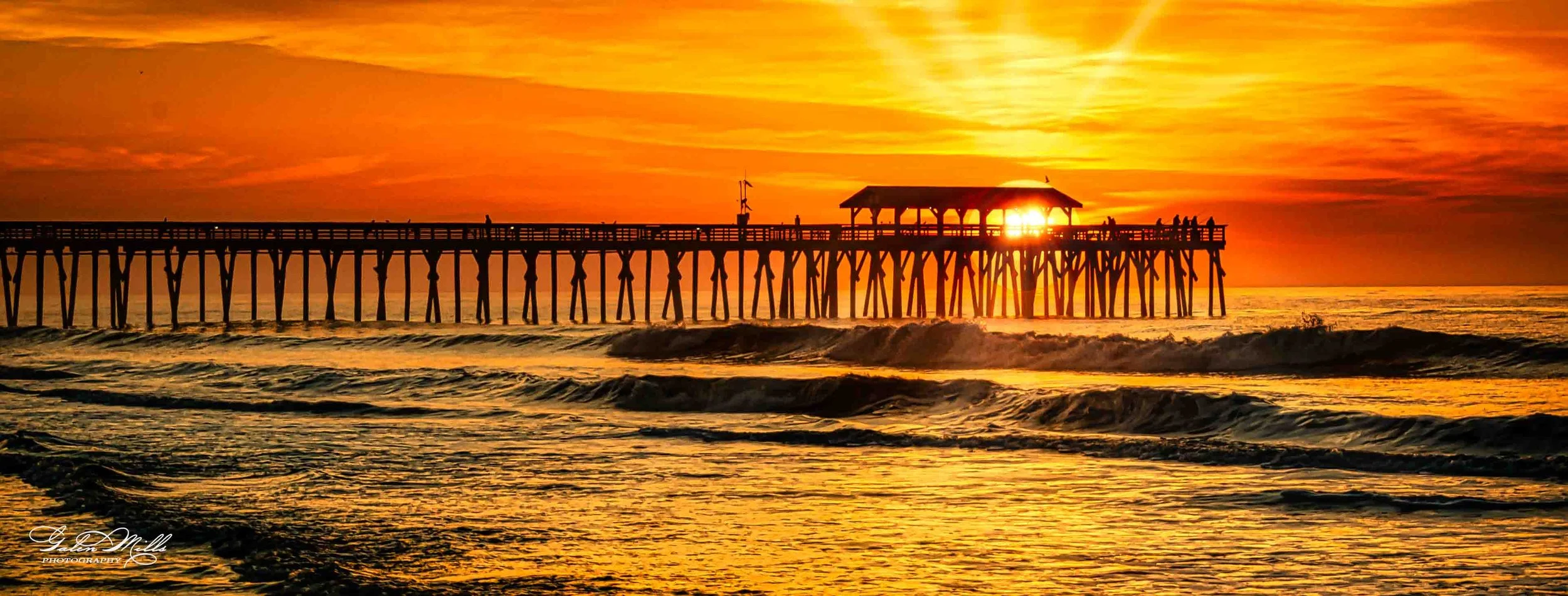 Sunset over the ocean with a pier extending into the water, silhouettes of people on the pier, and a small pavilion at the end, with the sun partially covered by the structure, creating a warm, golden glow.