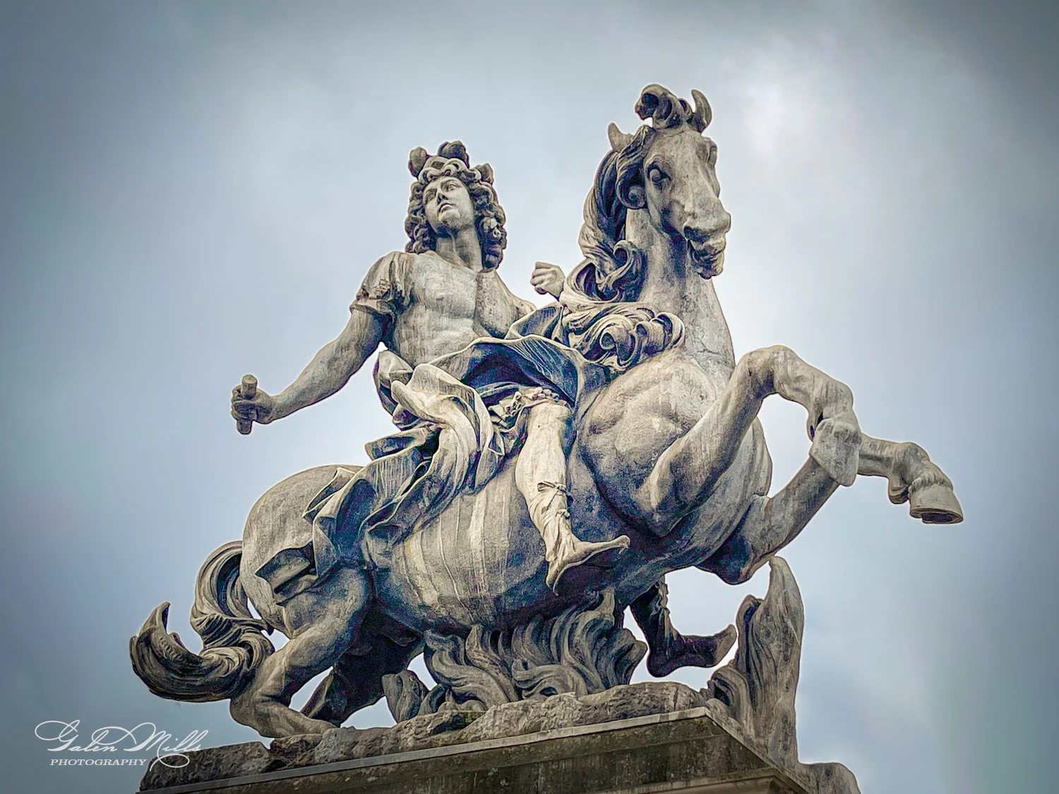 Statue of a man on a rearing horse, ornate details, gray stone, cloudy sky.