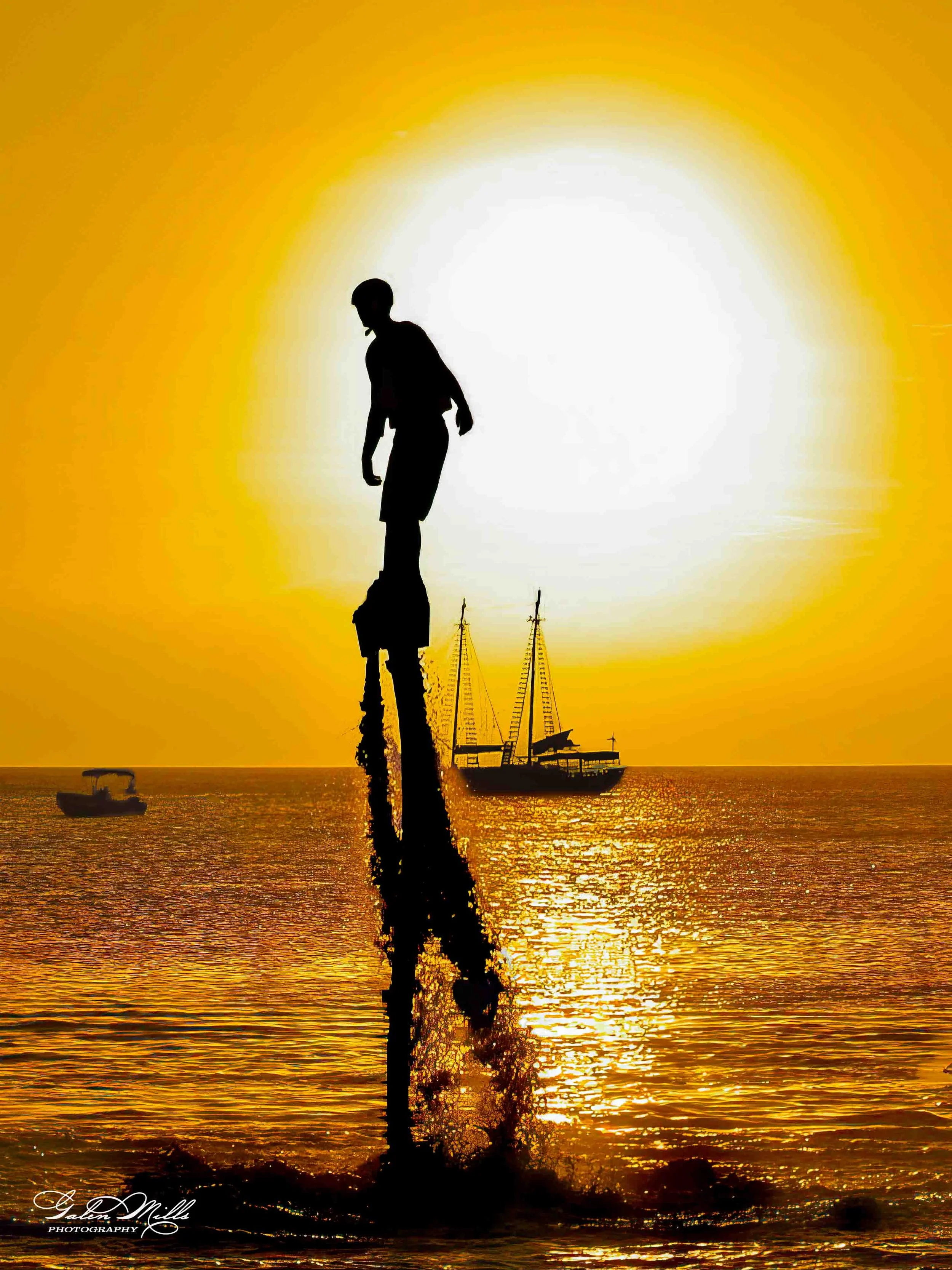 Silhouette of a person standing on top of a pole in water during sunset, with sailboats on the horizon.