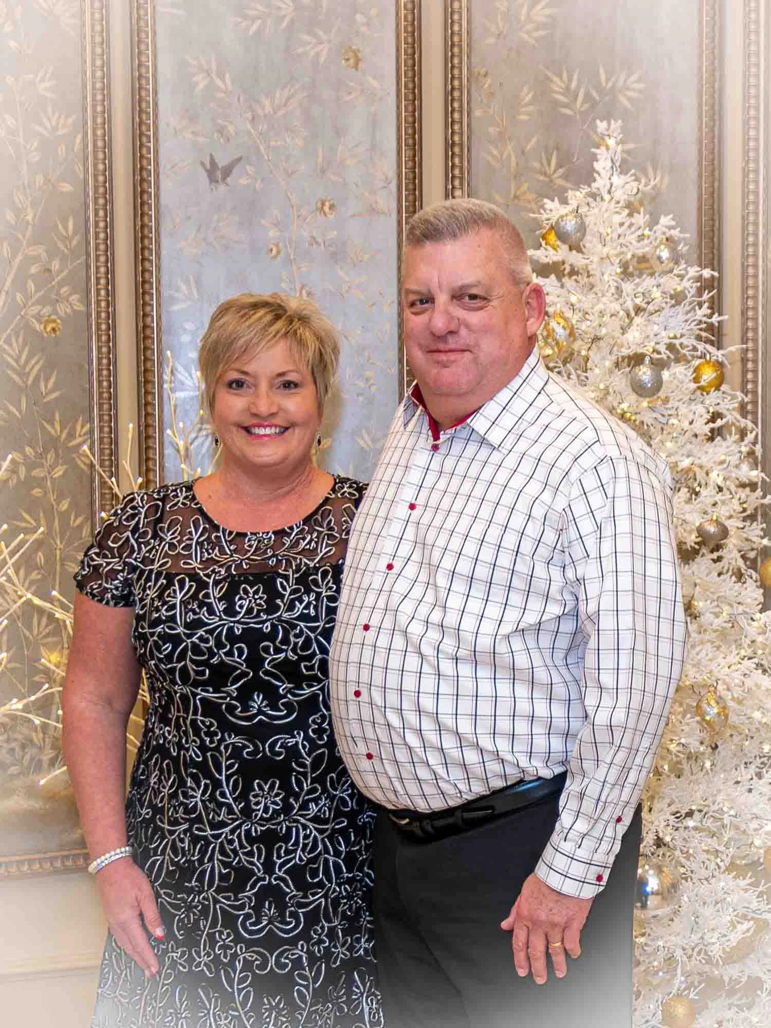 A smiling couple stands in front of a white Christmas tree with gold and silver ornaments, next to ornate wallpaper with floral designs.