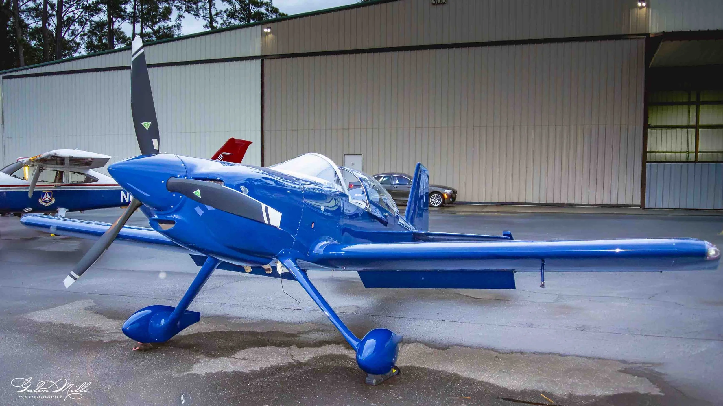 A blue single-engine propeller aircraft parked on the tarmac, with a larger airplane and a car in the background, outside a hangar.