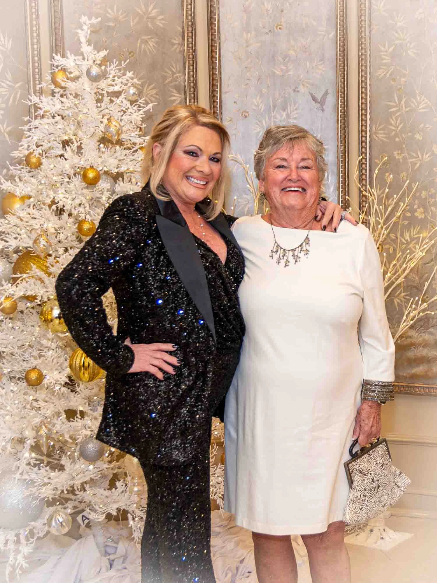 Two women smiling, standing in front of a white Christmas tree decorated with gold and silver ornaments. One woman is wearing a black sequin suit, and the other is in a white dress holding a clutch bag.