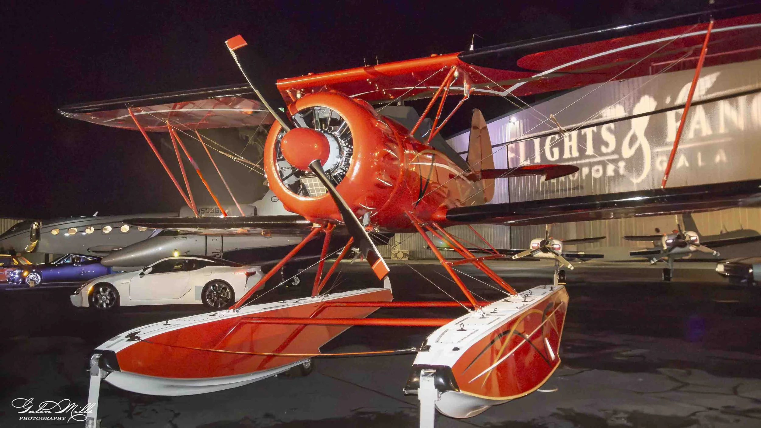 Red biplane on display at night, surrounded by cars, with a large illuminated sign in the background.