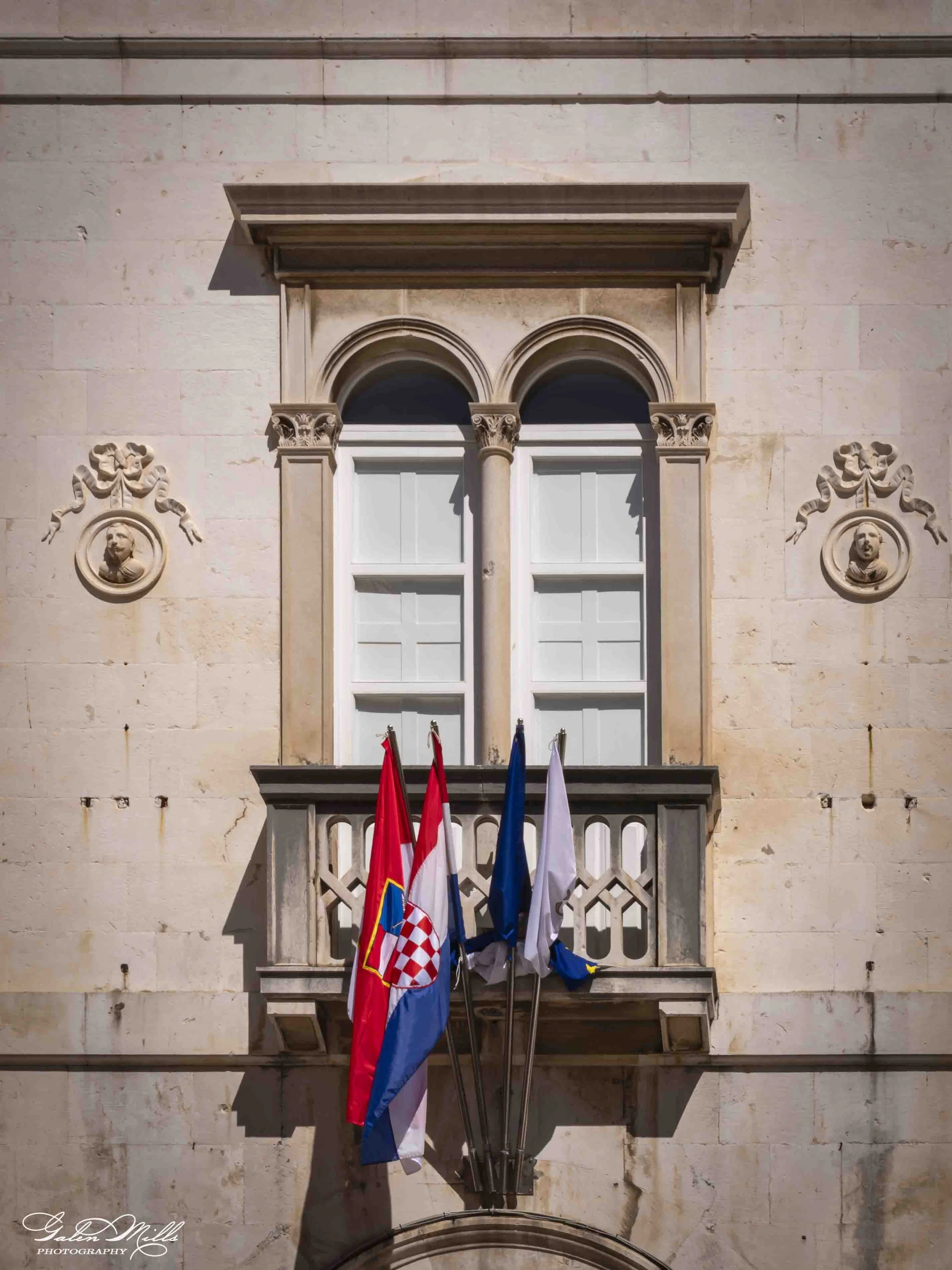 A European-style building facade with two arched windows, decorative columns, stone ornament carvings, and a small balcony with flags hanging below.