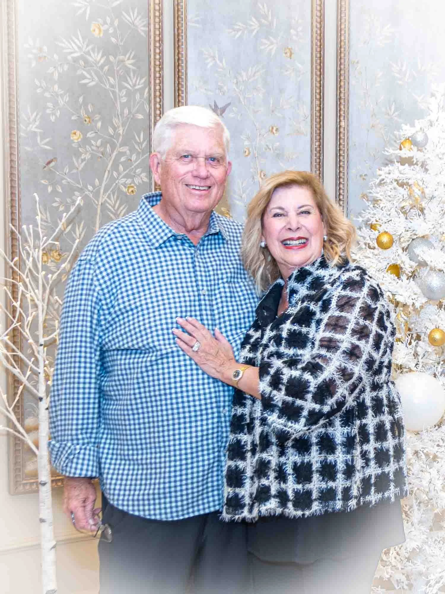 An older couple smiling and standing together in front of a decorative backdrop with white and gold elements, including a white-tipped artificial tree with gold ornaments. The man is wearing a blue checkered shirt, and the woman is wearing a black-an