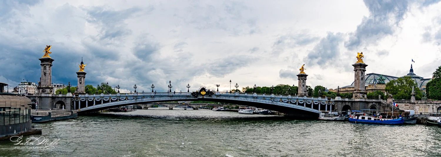Pont Alexandre III bridge in Paris with ornate sculptures and gilded statues, spanning the Seine River, surrounded by boats and classic architecture under a cloudy sky.