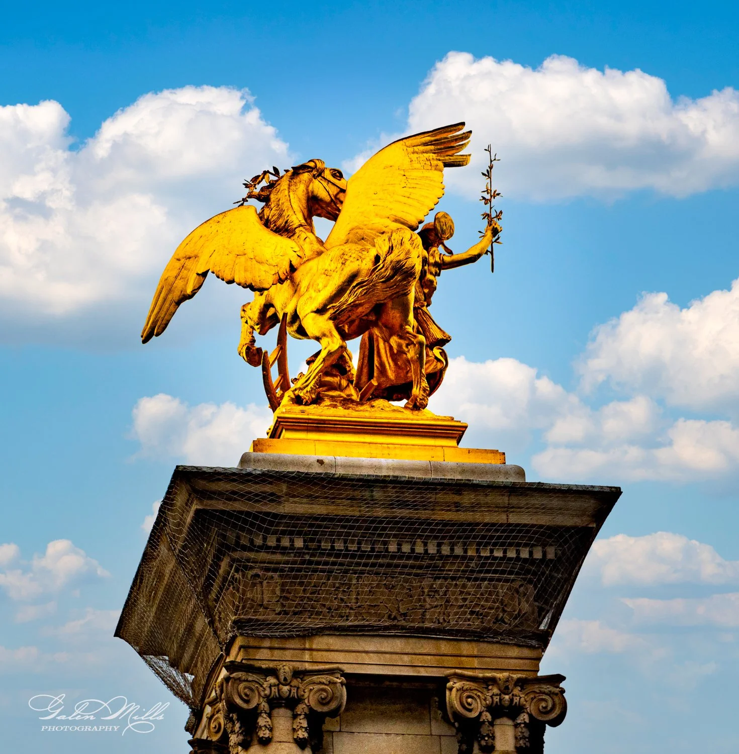 Golden statue of a winged horse with a rider holding an olive branch, set on a stone pedestal against a blue sky with clouds.
