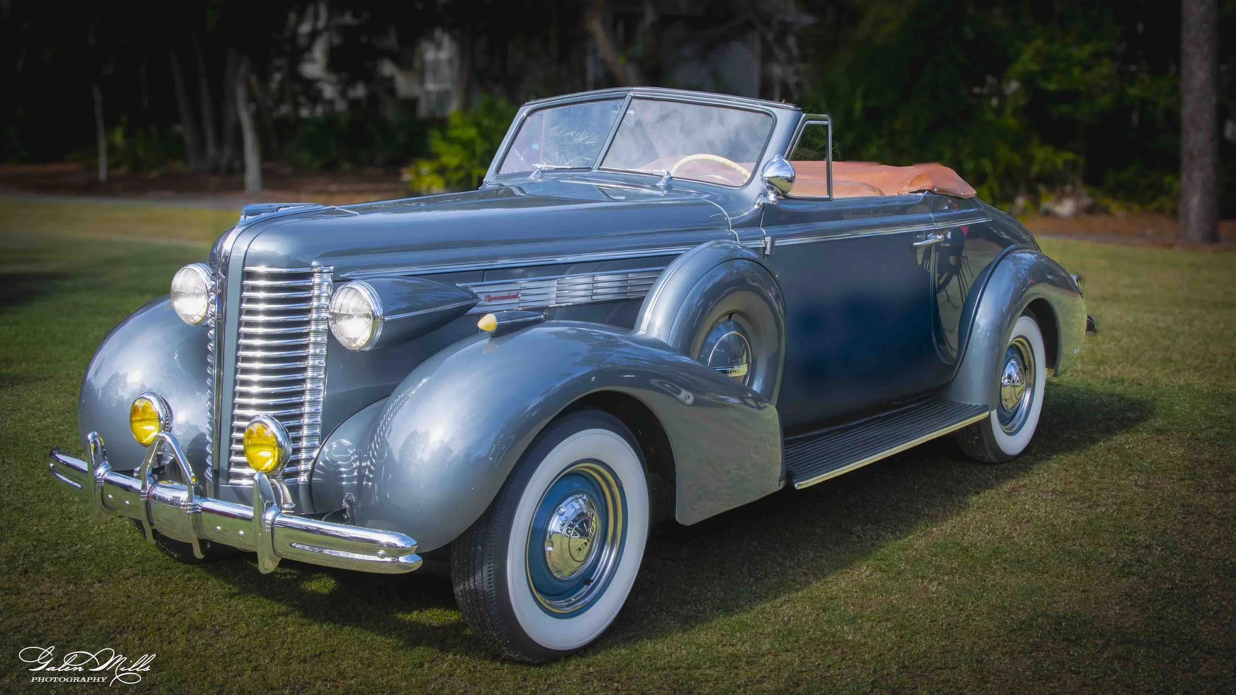A vintage blue convertible car with whitewall tires parked on a grassy area, with trees and greenery in the background.