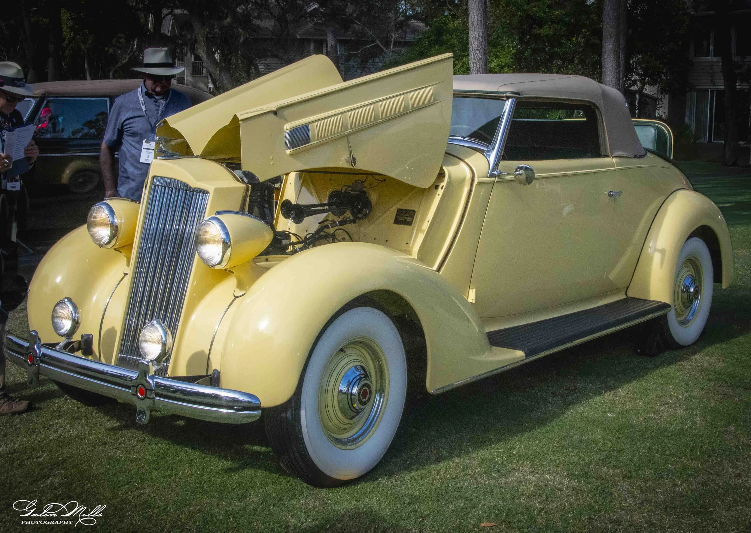 A vintage yellow convertible car with the hood open, displaying its engine, parked on grass at an outdoor event. Several people are nearby, some with hats and event badges, observing the car.