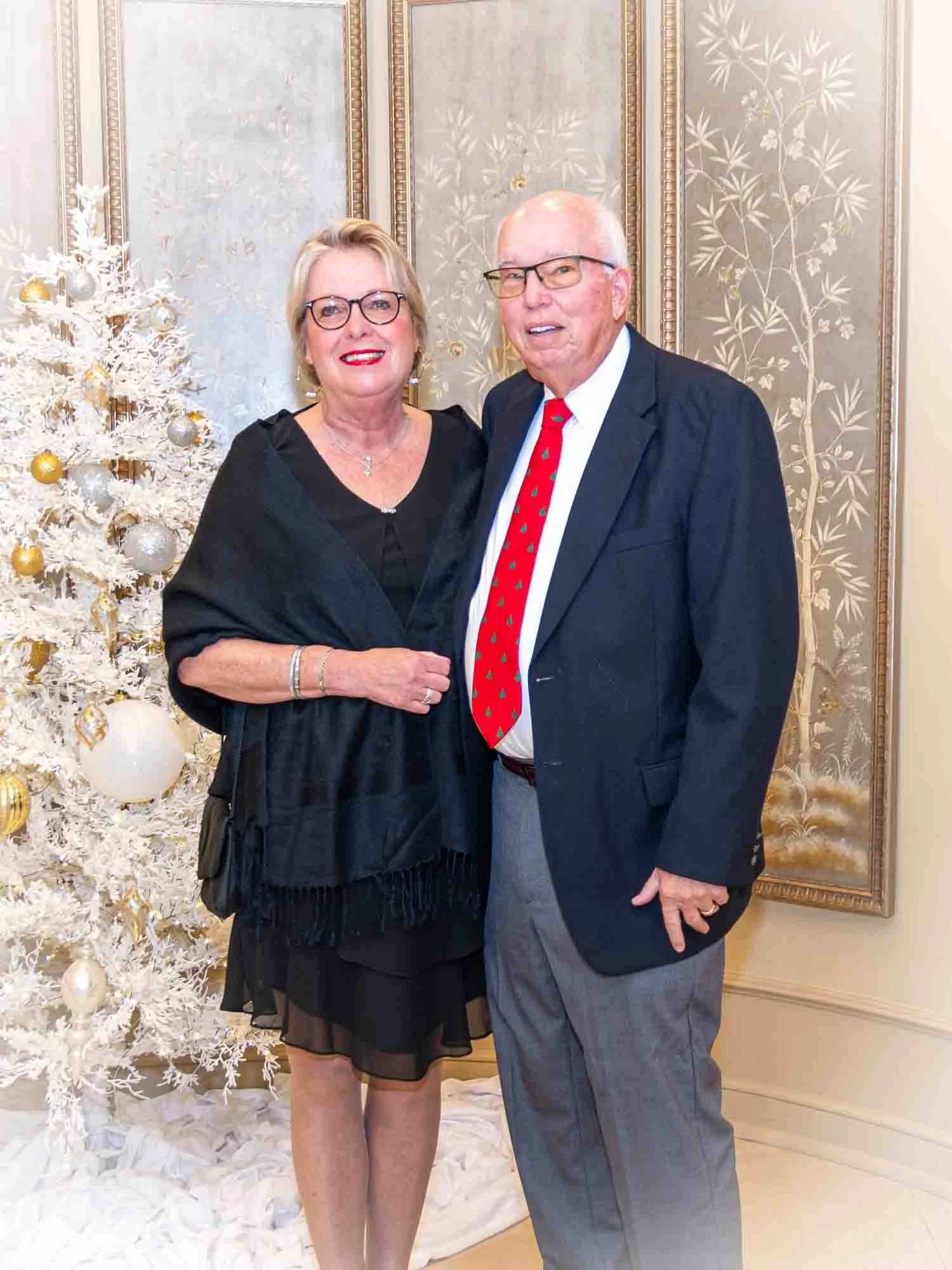A smiling couple posing in front of a decorated white Christmas tree, wearing formal attire. The woman is in a black dress and the man is in a suit with a red tie. The background has ornate paneling.