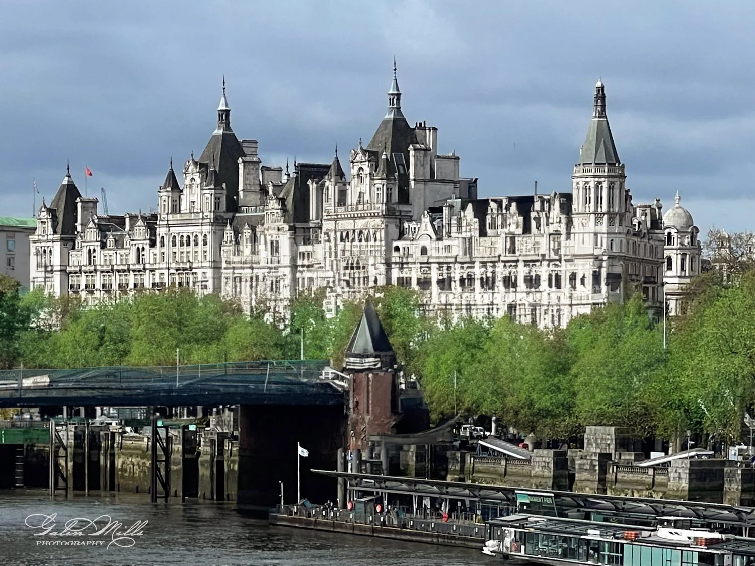 Large historic building with Gothic architecture along a river, surrounded by green trees and boats docked by the shore.