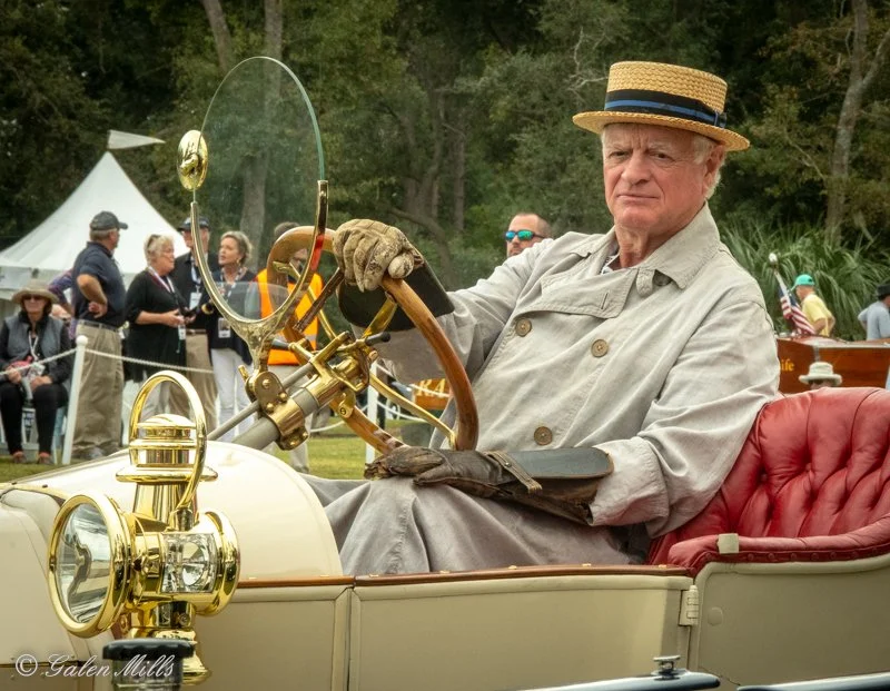 Elderly man in vintage attire driving an antique car at an outdoor event with spectators in the background.