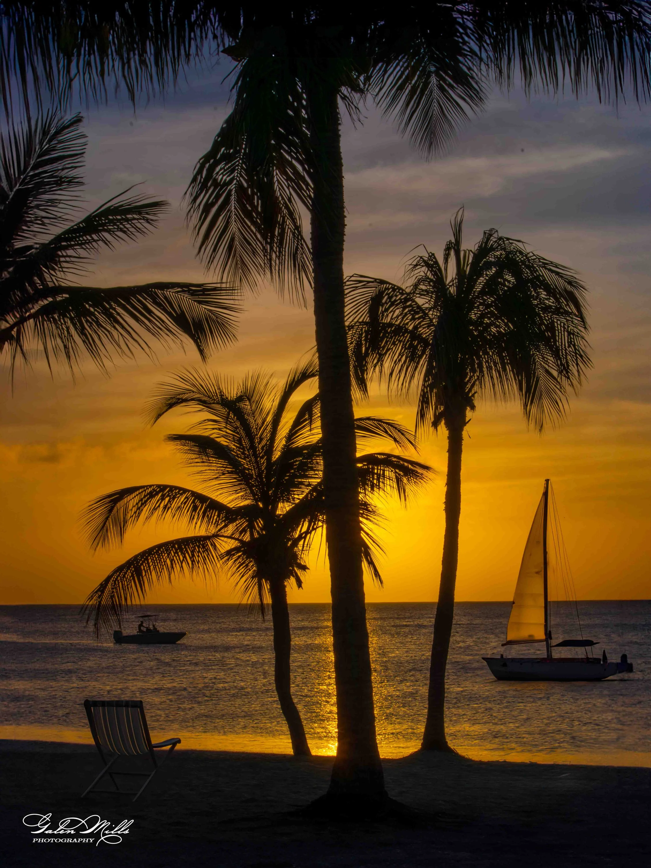 A tropical beach at sunset with silhouettes of palm trees, a sailboat, and a small motorboat on calm water, and a single beach chair on the sand.