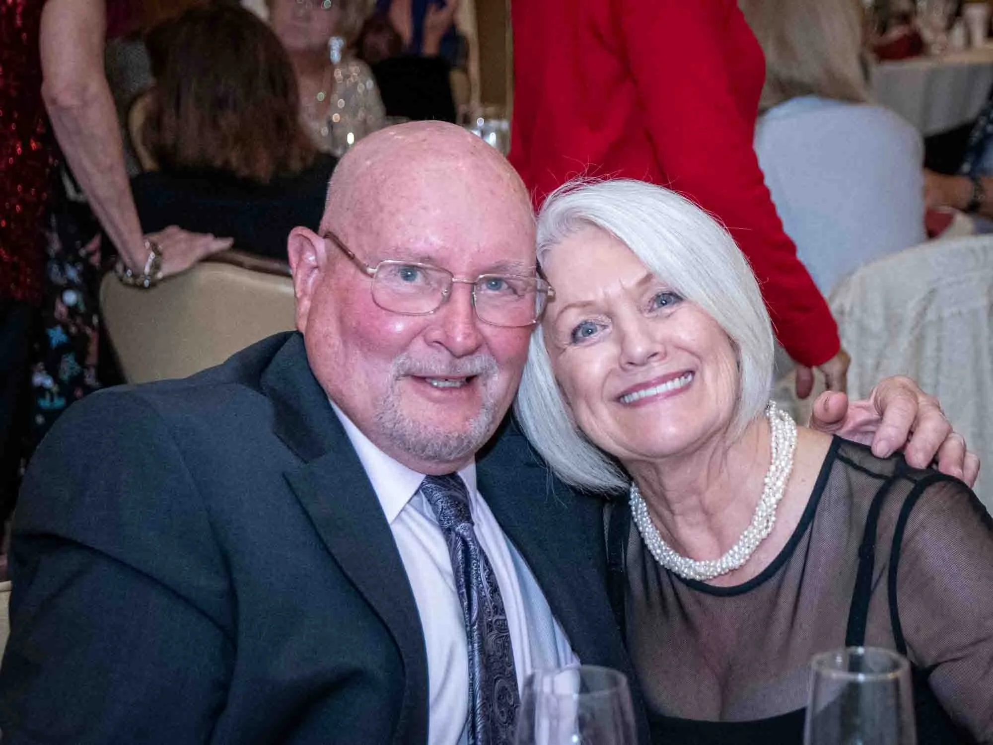 A smiling man and woman at an event sitting close together. The man is wearing glasses, a suit, and tie. The woman has short white hair, wearing a black dress and pearl necklace. Background shows more people seated at tables.