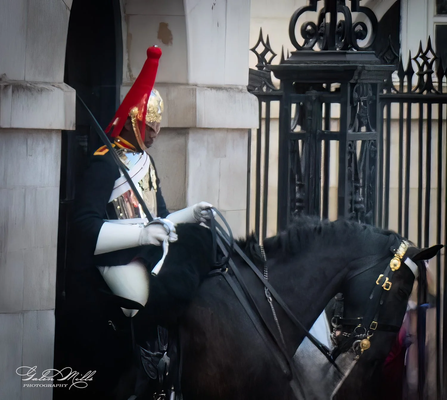 British cavalry guard in ceremonial uniform and red helmet seated on a black horse in front of a stone archway and metal gate.