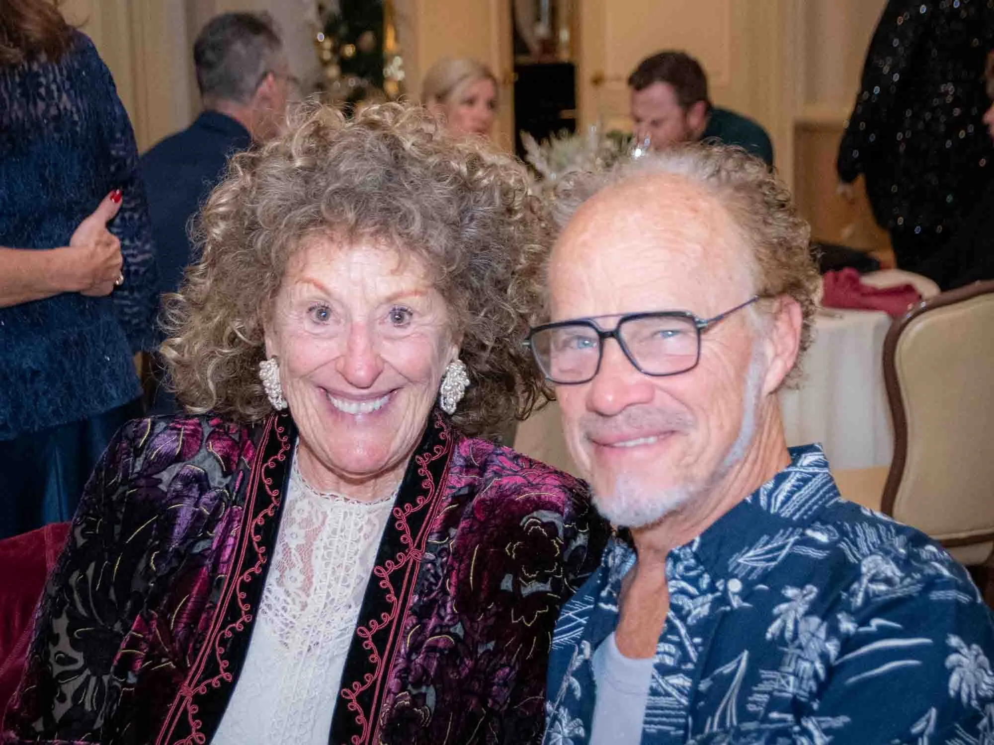 A smiling elderly couple sitting together at a social event, surrounded by other guests. The woman has curly hair and is wearing ornate earrings and a colorful jacket. The man is wearing glasses, has short hair, and is in a patterned shirt. Tables an