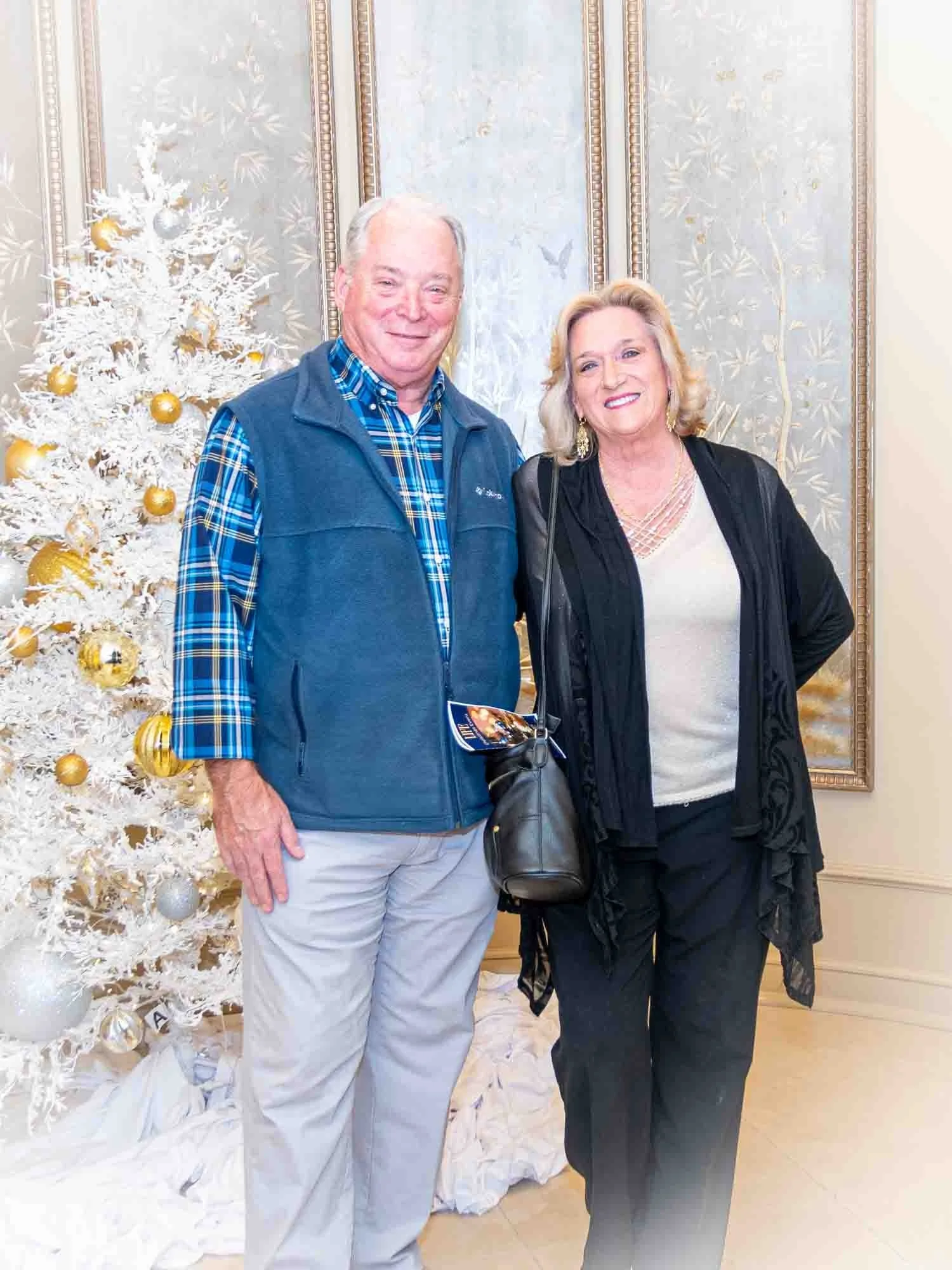 A man and woman posing in front of a white decorated Christmas tree with gold and silver ornaments, standing in an elegantly decorated room.