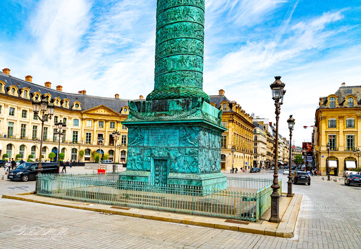 A large green column with intricate carvings in a European city square, surrounded by historic buildings, streetlights, and parked cars.