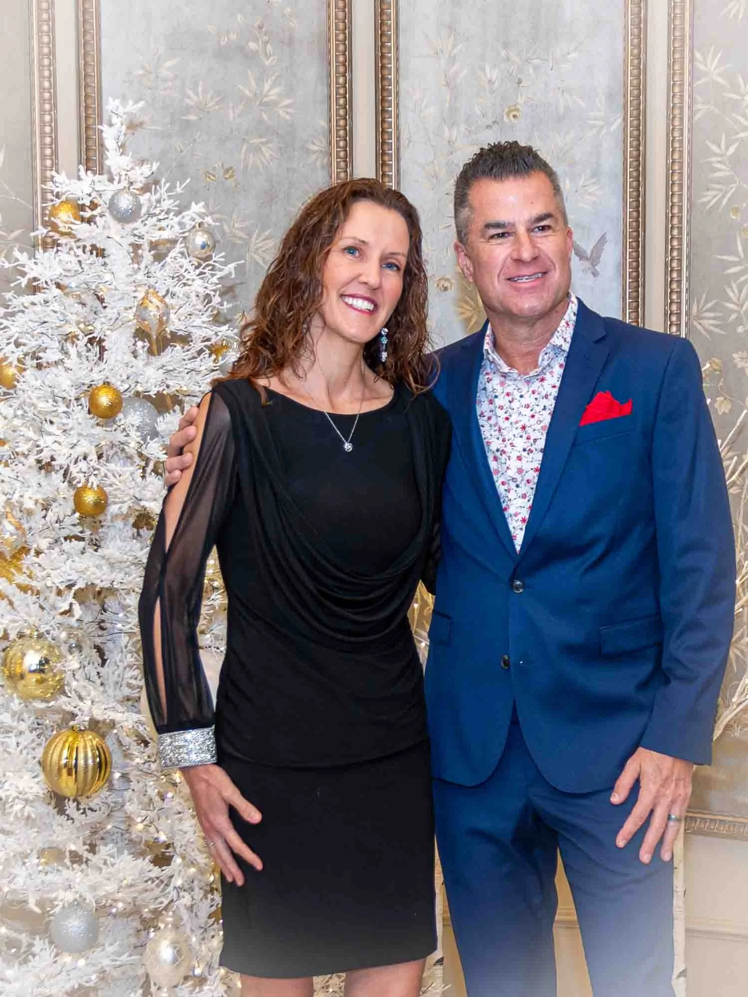 A couple posing in formal attire next to a white Christmas tree with gold and silver ornaments.