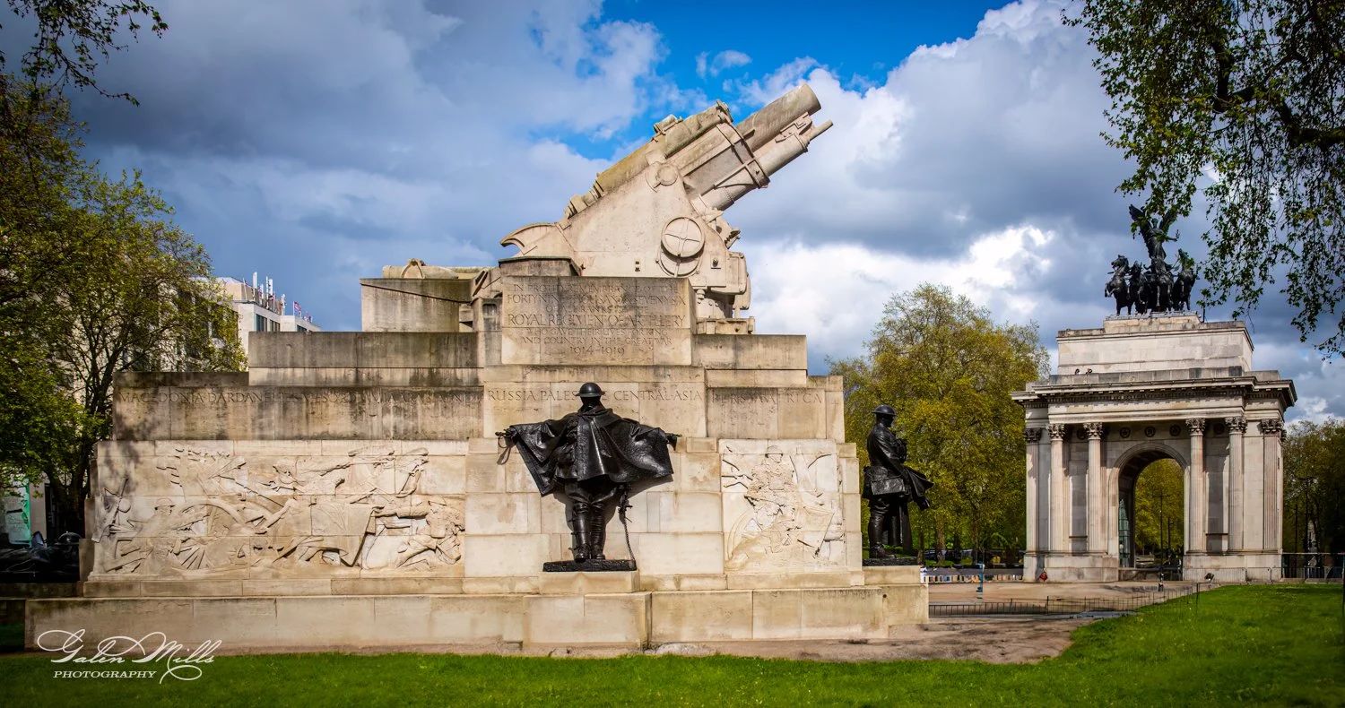 War memorial with artillery sculpture and statue, near a triumphal arch with a horse-drawn chariot sculpture on top, surrounded by trees and grass under a cloudy sky.