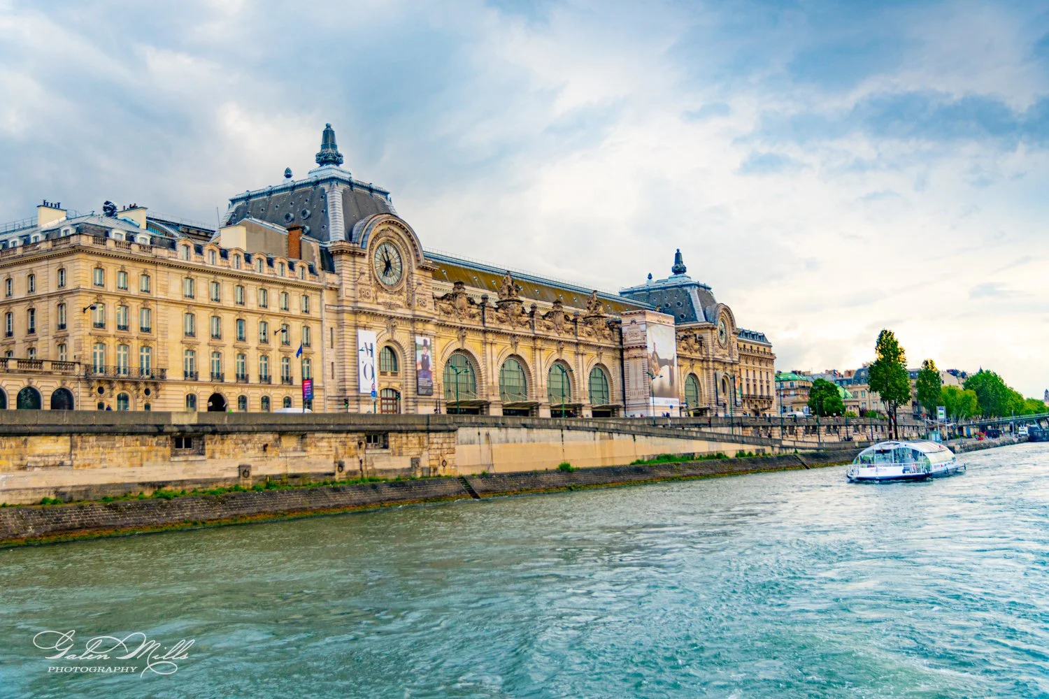 Historic stone building by a river with a clock tower, and a boat on the water under a cloudy sky.
