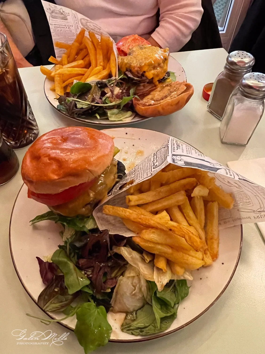 Two plates of burgers with cheese, lettuce, tomato, and buns, served with side dishes of French fries and leafy green salad. A soft drink and salt and pepper shakers are on the table.