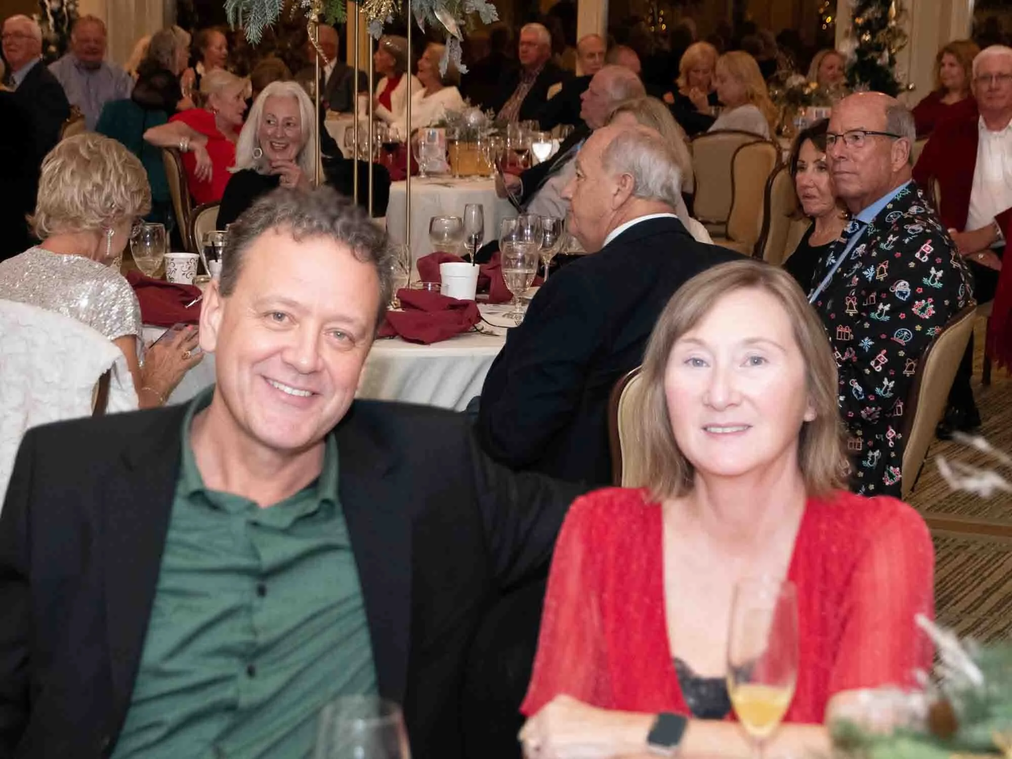 A man in a green shirt and black jacket sits next to a woman in a red dress at a formal event. They are seated at a table with glasses and a centerpiece in a crowded banquet hall.