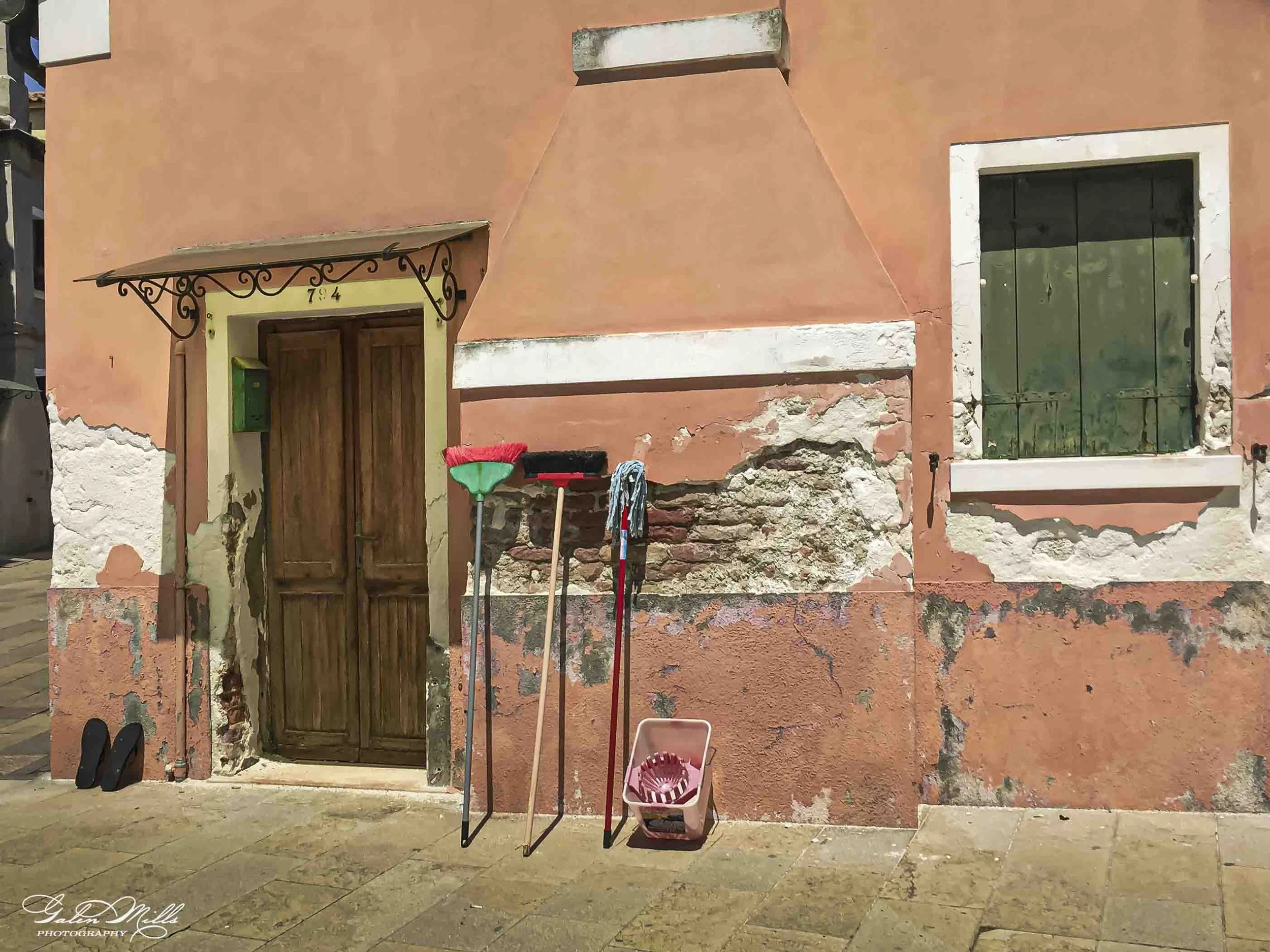 Outdoor scene with a weathered pinkish-orange stucco wall, a wooden door under a decorative metal awning, and a window with green shutters. Various cleaning tools and a pink basket are leaning against the wall, with shoes on the ground nearby.