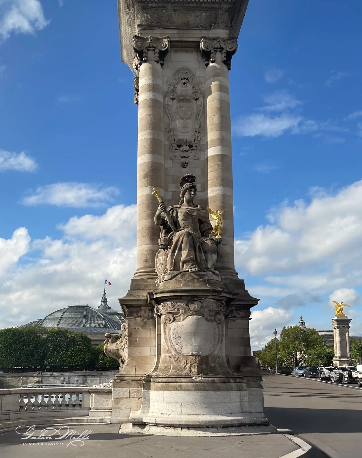 Stone statue and column on Pont Alexandre III, Paris, with blue sky and dome in background.