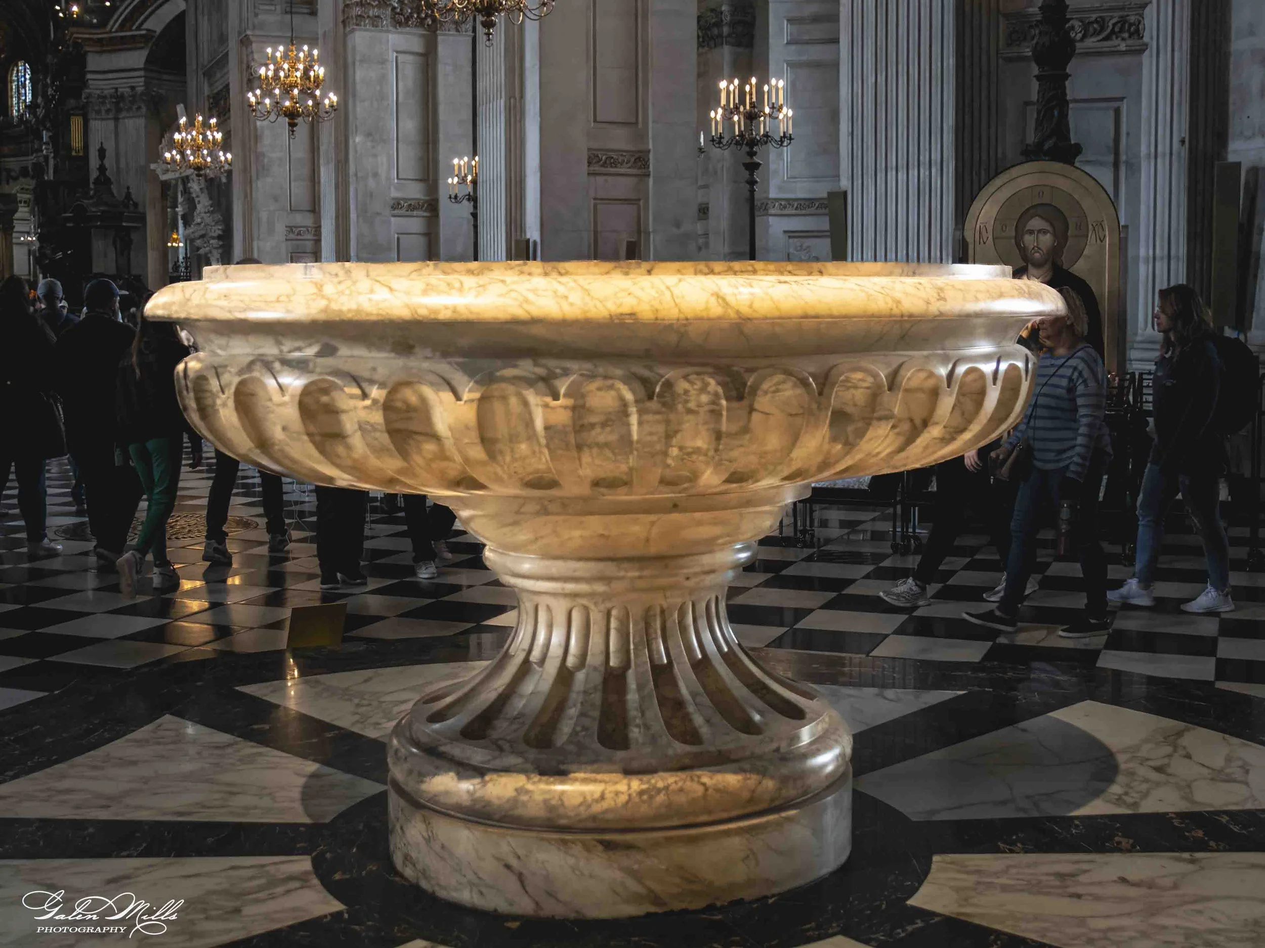 A large marble baptismal font in a church, surrounded by chandeliers and tall columns with people nearby.