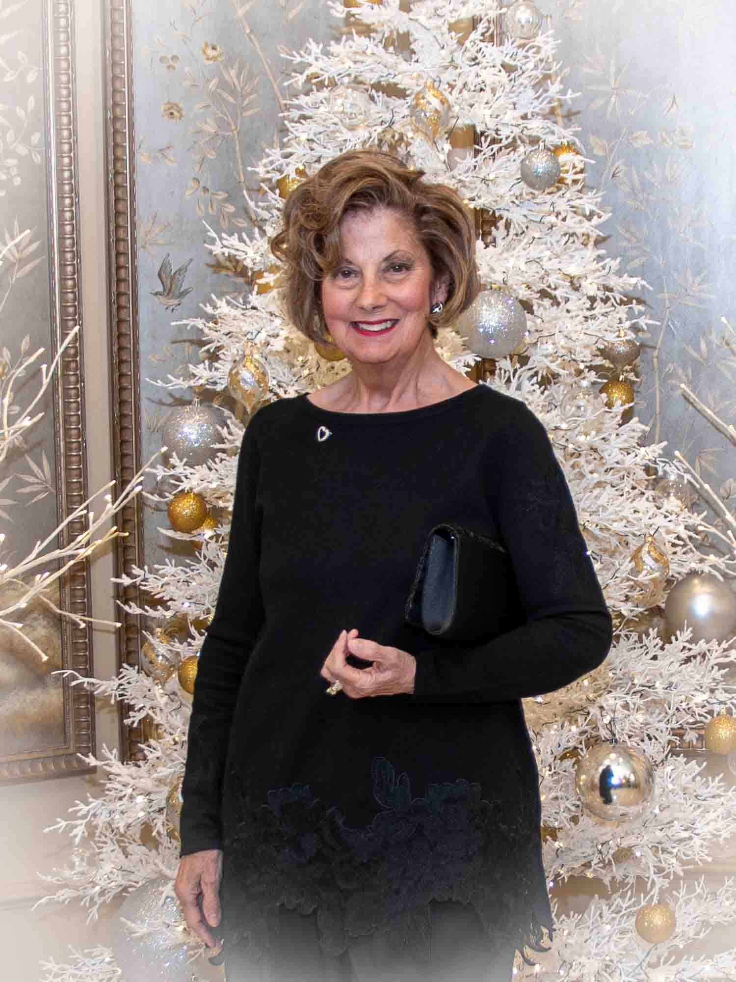A woman in a black outfit stands in front of a white Christmas tree decorated with gold and silver ornaments.