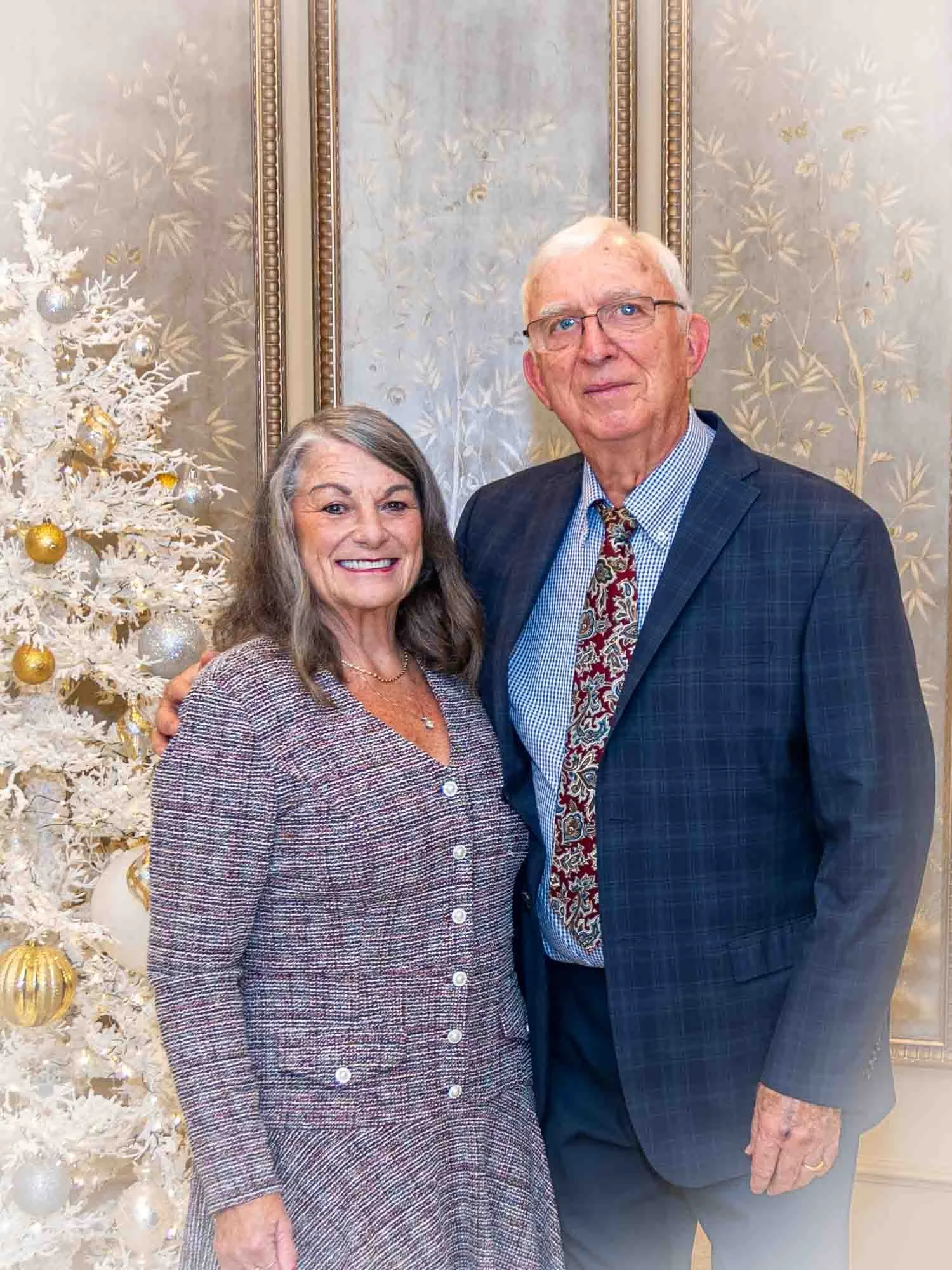 An older couple poses in front of a decorated Christmas tree, with a festive background. The woman is wearing a patterned suit, and the man is in a suit with a patterned tie. They are smiling.