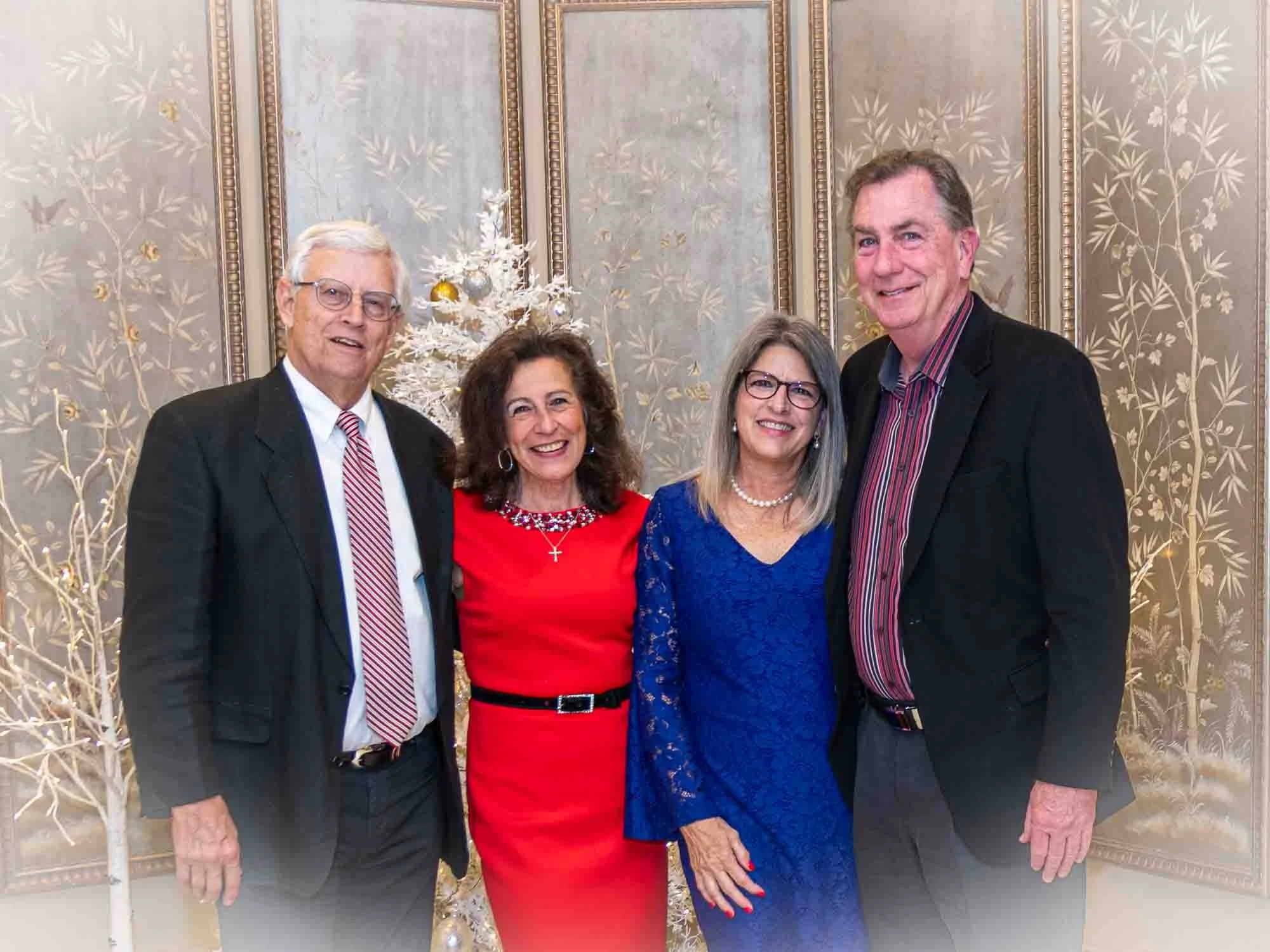 Four people smiling in formal attire in front of ornate wallpaper and a decorative white tree, with two women in red and blue dresses and two men in suits.