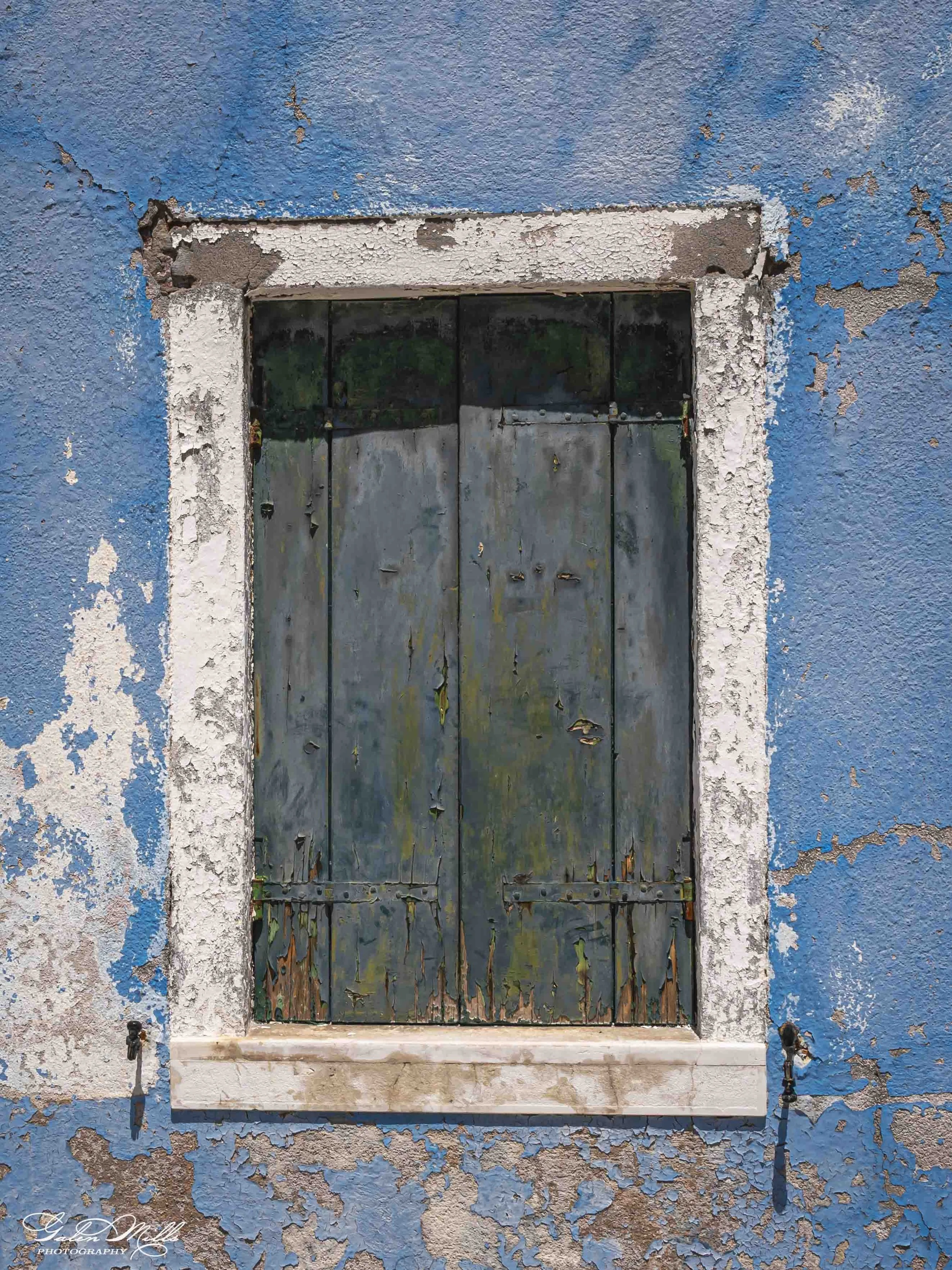 Old, weathered window with peeling paint on a vibrant blue wall.