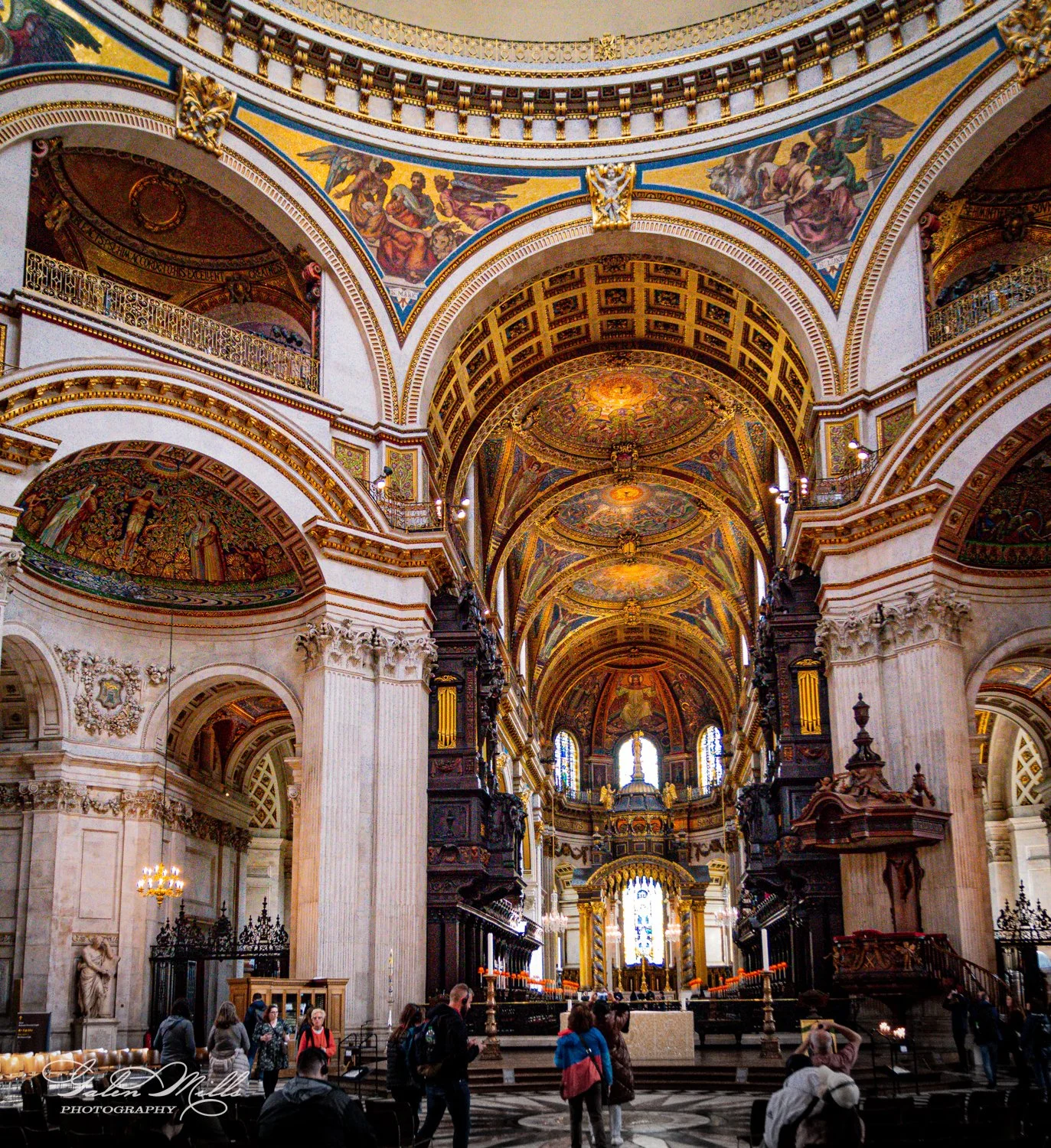 Interior of a grand cathedral with ornate ceiling, decorative arches, and large columns, featuring intricate artwork and stained glass windows. People are walking inside the cathedral.