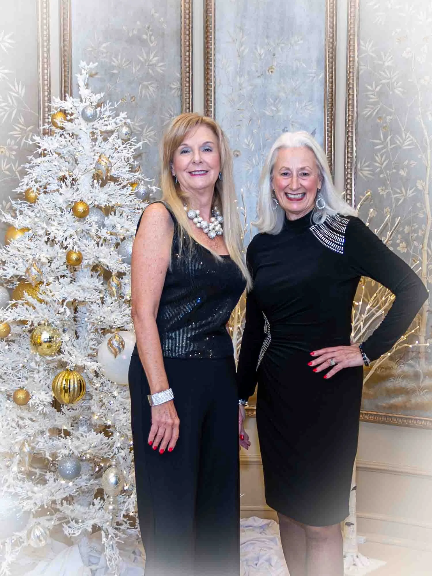 Two women in elegant black outfits standing next to a decorated white Christmas tree with gold and silver ornaments.