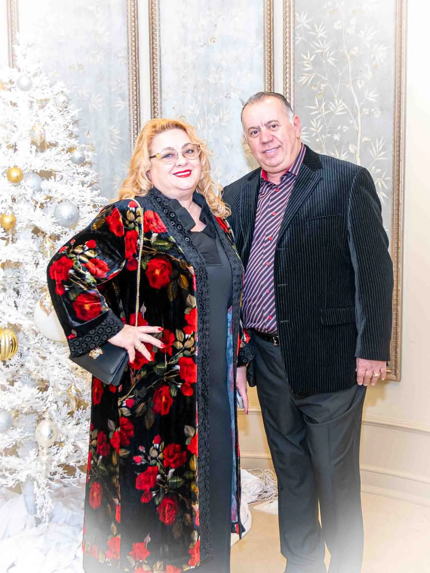 A woman in a floral print coat and a man in a striped suit standing next to a white Christmas tree with gold and silver ornaments.