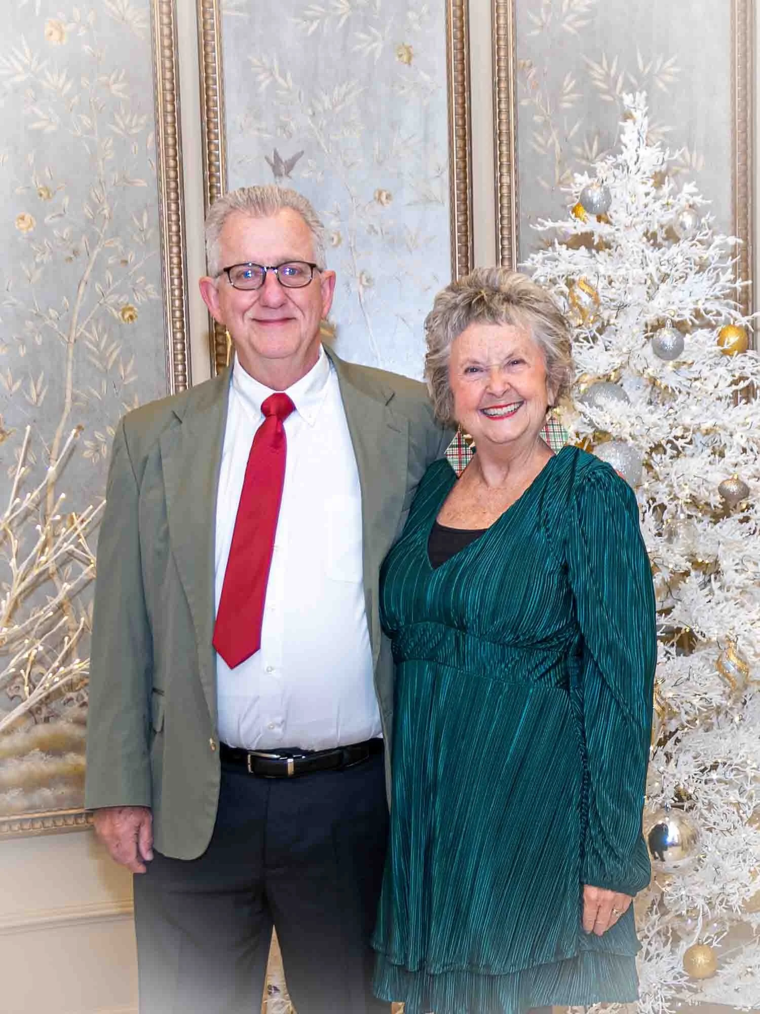 An older couple standing together in front of a decorated white Christmas tree with gold and silver ornaments. The man is wearing a suit with a red tie, and the woman is in a green dress. They are smiling at the camera in a festive, elegant setting.