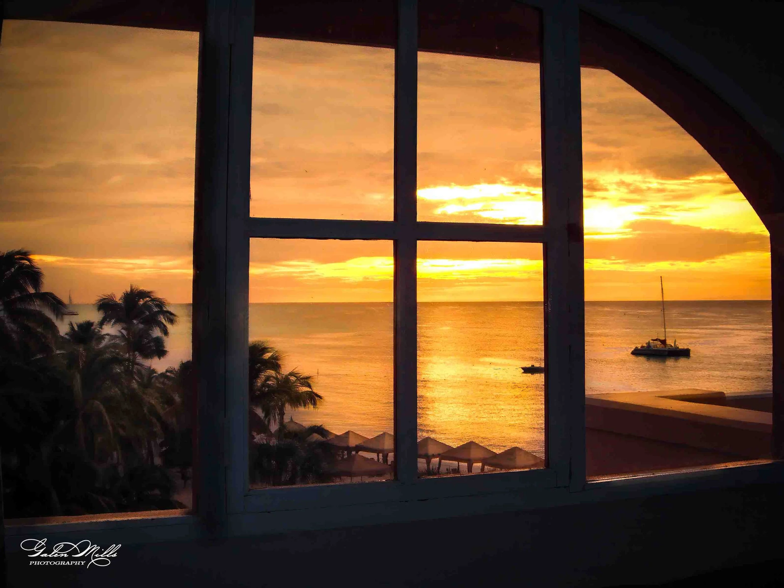 A sunset view of the ocean through a window with multiple panes. Palm trees and beach umbrellas are visible in the foreground, and a sailboat is on the water.