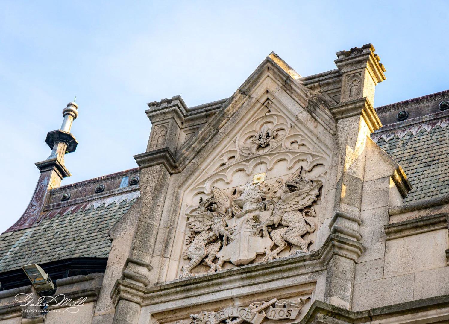 Gothic architectural detail with intricate carvings and a coat of arms featuring two griffins, set against a background of stonework and a blue sky.