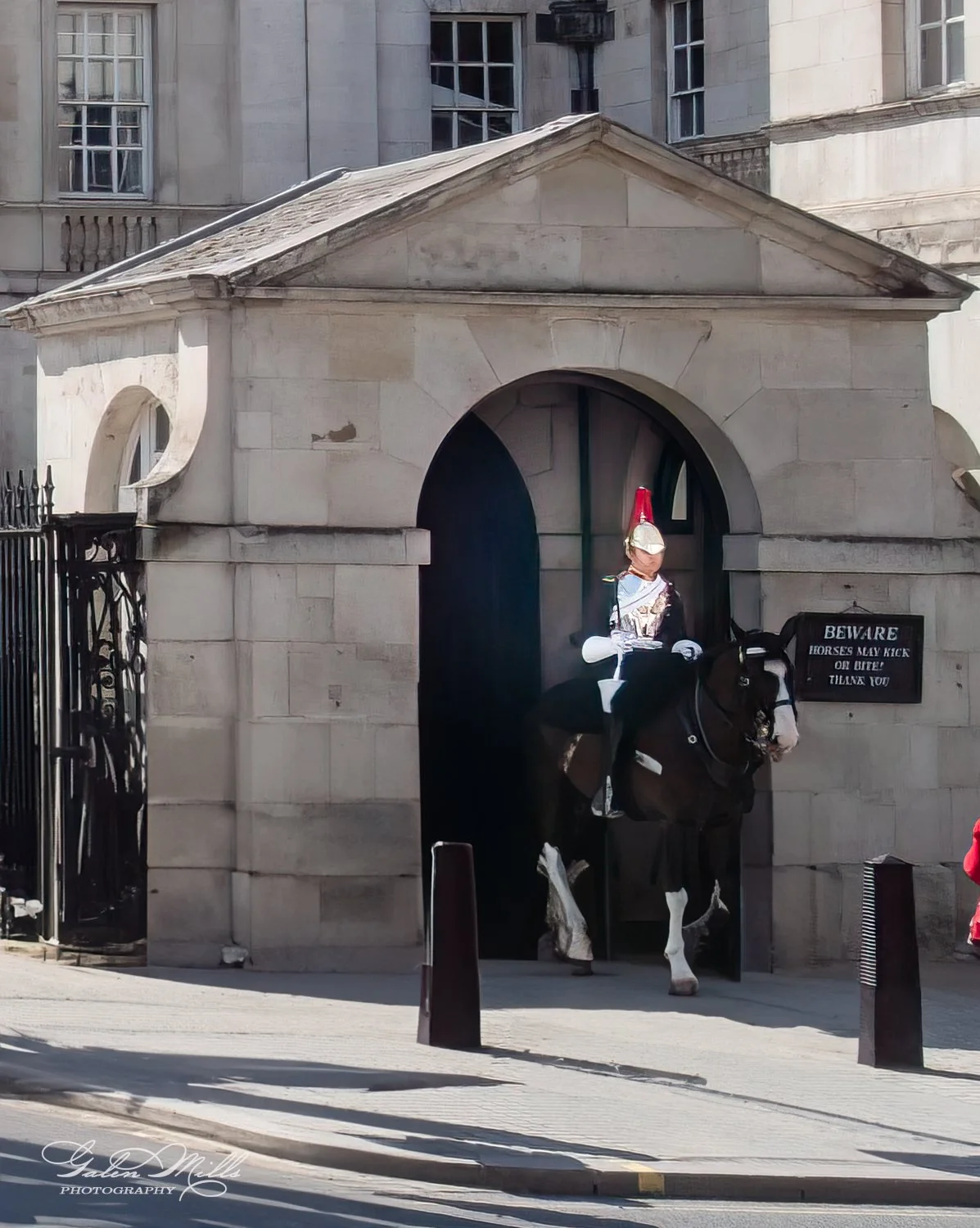 A mounted guard in traditional uniform on horseback exiting a stone building with an arched entrance. A sign next to the entrance warns about horses kicking or biting.