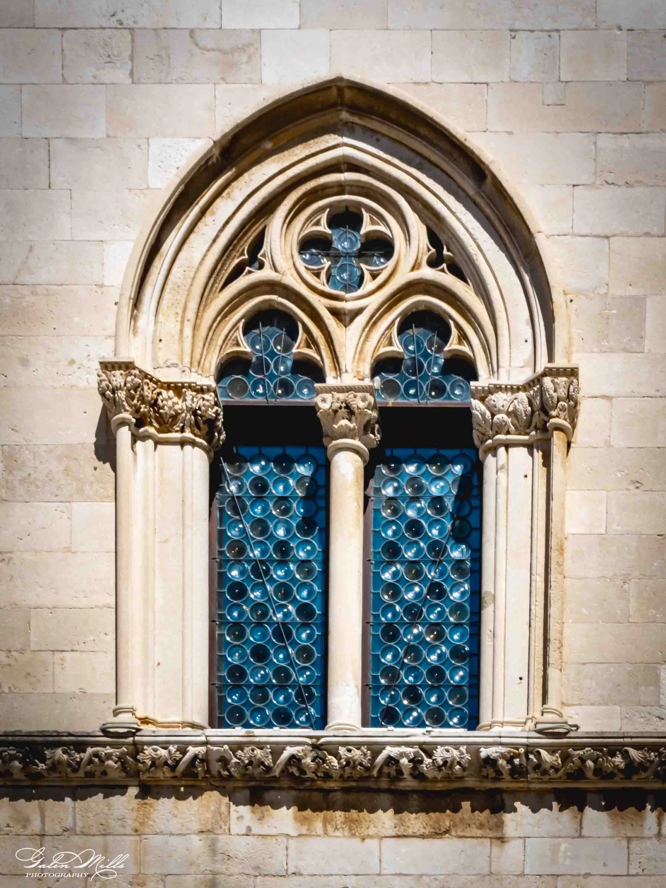Close-up of a Gothic-style stone window with stained glass, featuring blue and clear circular glass pieces, ornate carved stone tracery, and decorative sculpted details on the window frame.