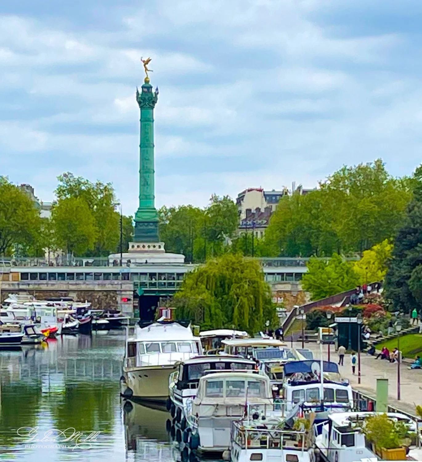 View of the July Column with boats docked on a canal in Paris, surrounded by trees and buildings under a cloudy sky.