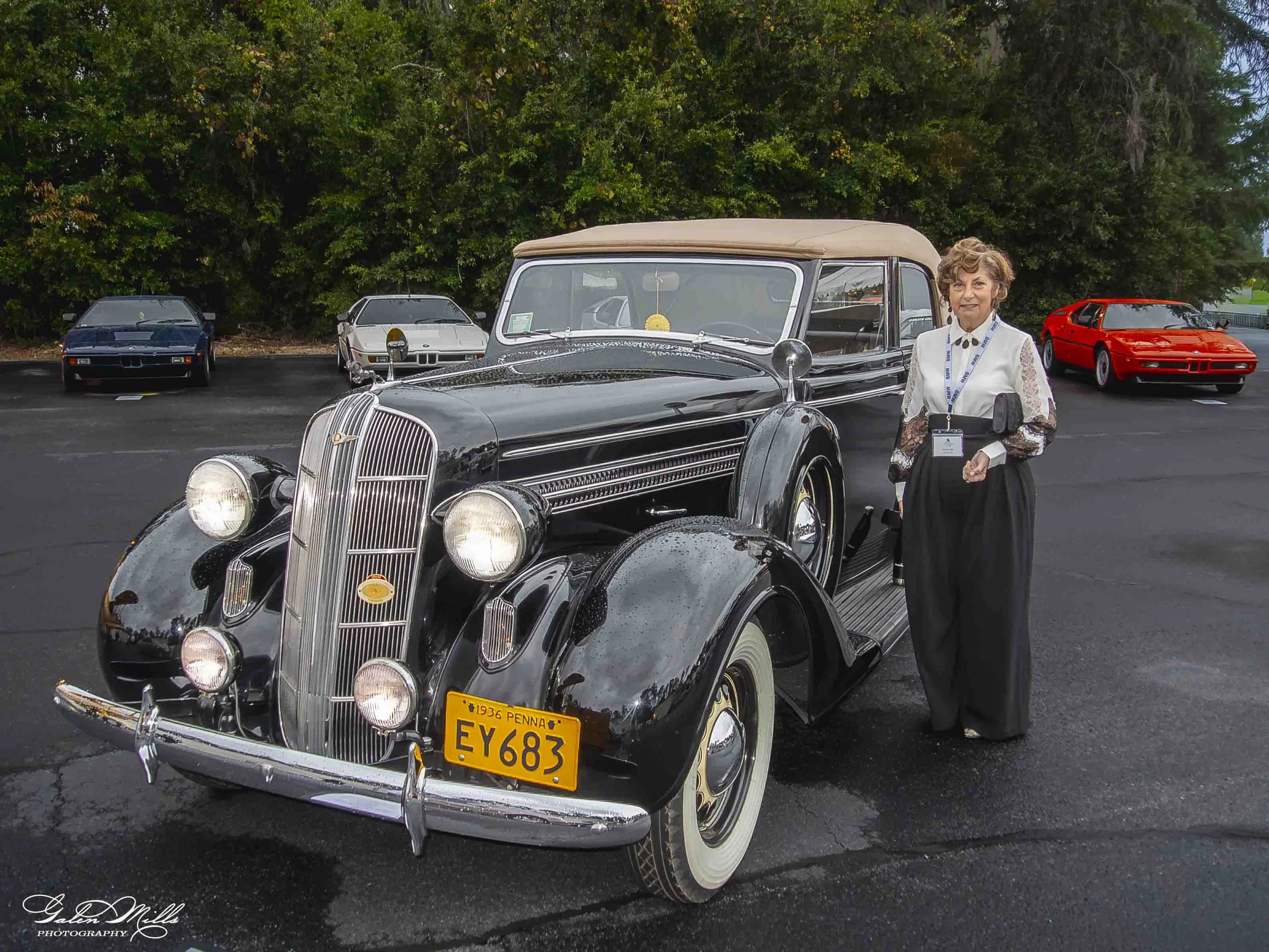 A black vintage 1936 car with a beige roof, parked outdoors with three modern cars and lush green trees in the background. A woman with short curly hair, wearing a white lace top and a badge, stands beside the vintage car.