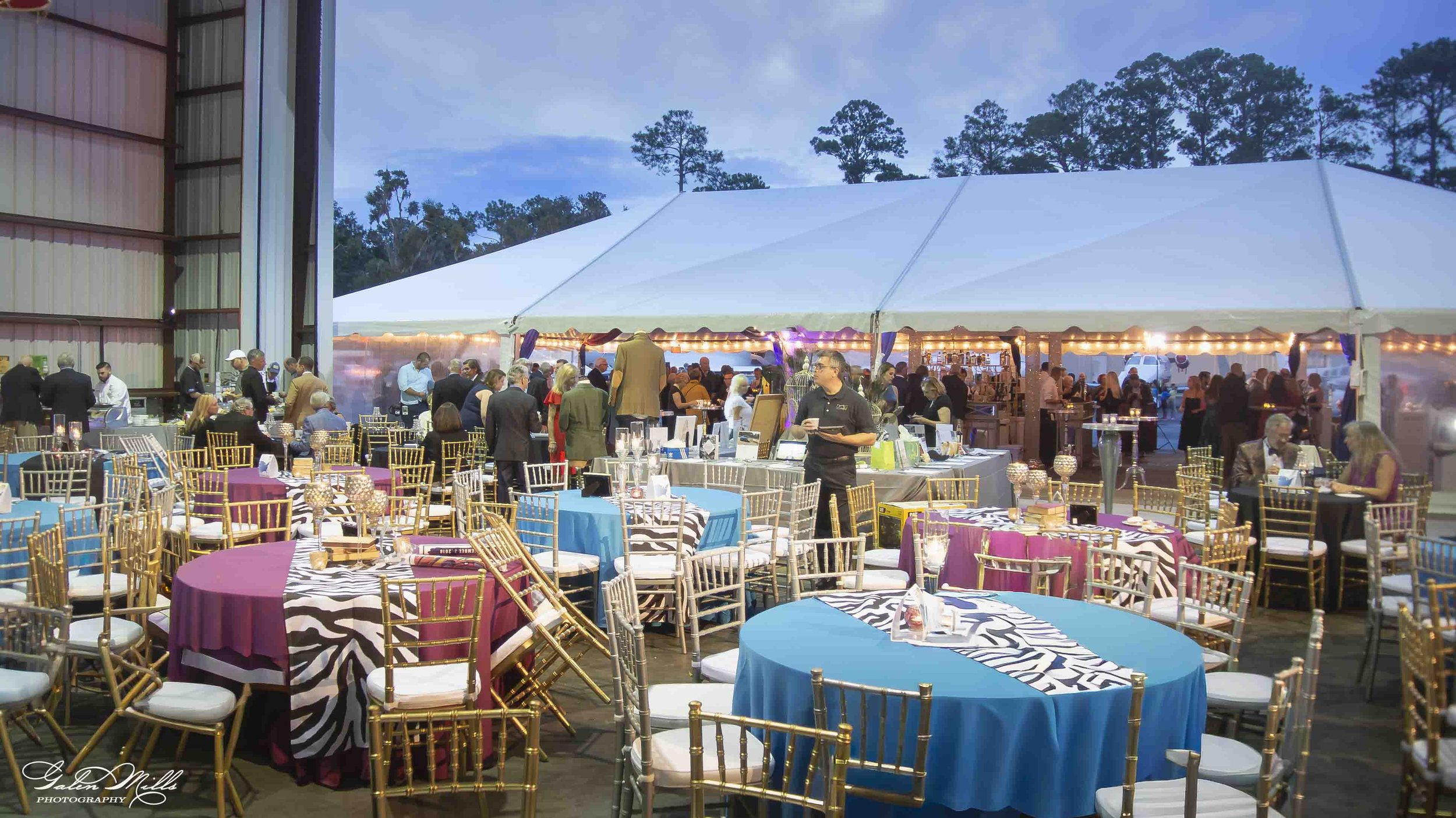 Indoor event space with round tables covered in colorful tablecloths and gold chairs, filled with people mingling and dining under a large white tent, with outdoor trees visible in the background, during dusk or early evening.