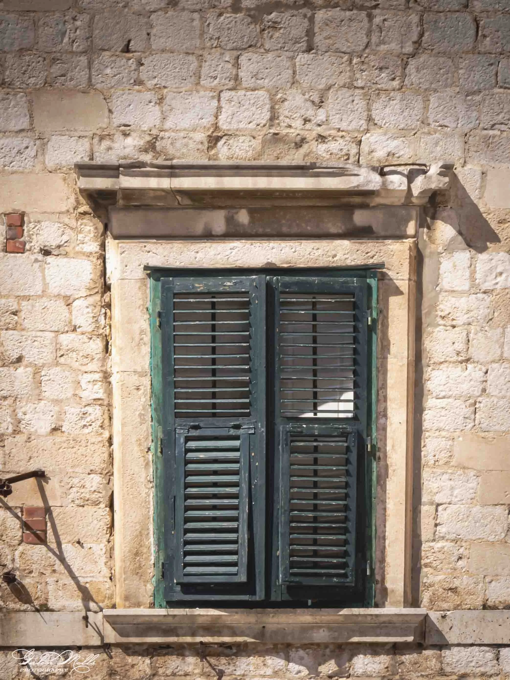 A stone building with green window shutters and a decorative stone lintel above the window. The building has a textured exterior with bricks and mortar.