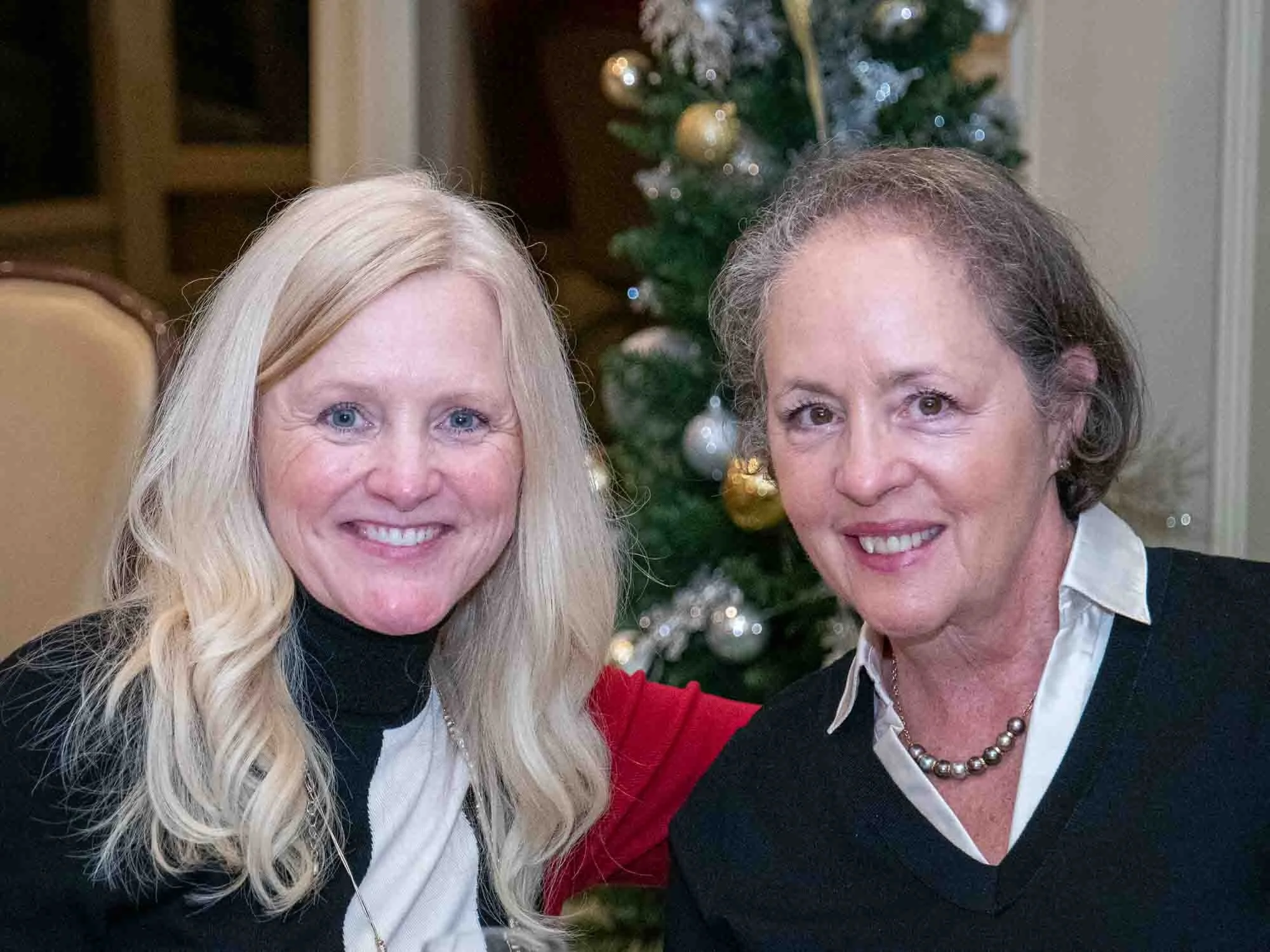 Two women smiling in front of a decorated Christmas tree indoors.
